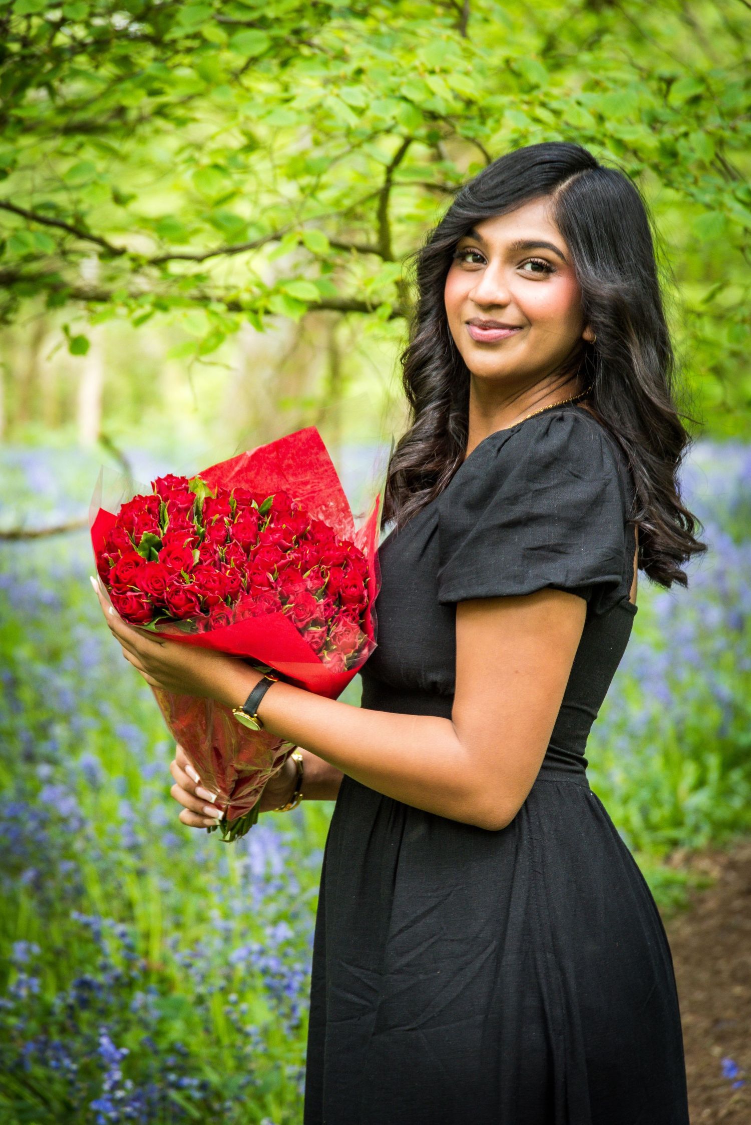 Woman in black dress holding a bouquet of red roses in a bluebell woodland.