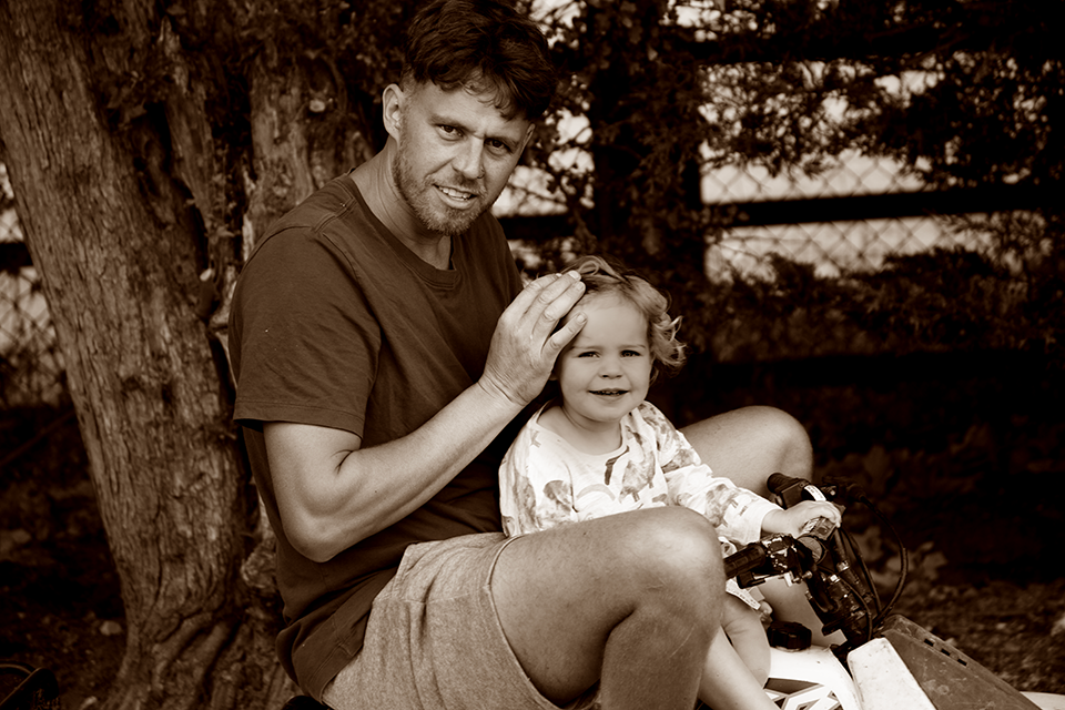 Sepia toned portrait of father sitting under tree with young daughter on his lap.