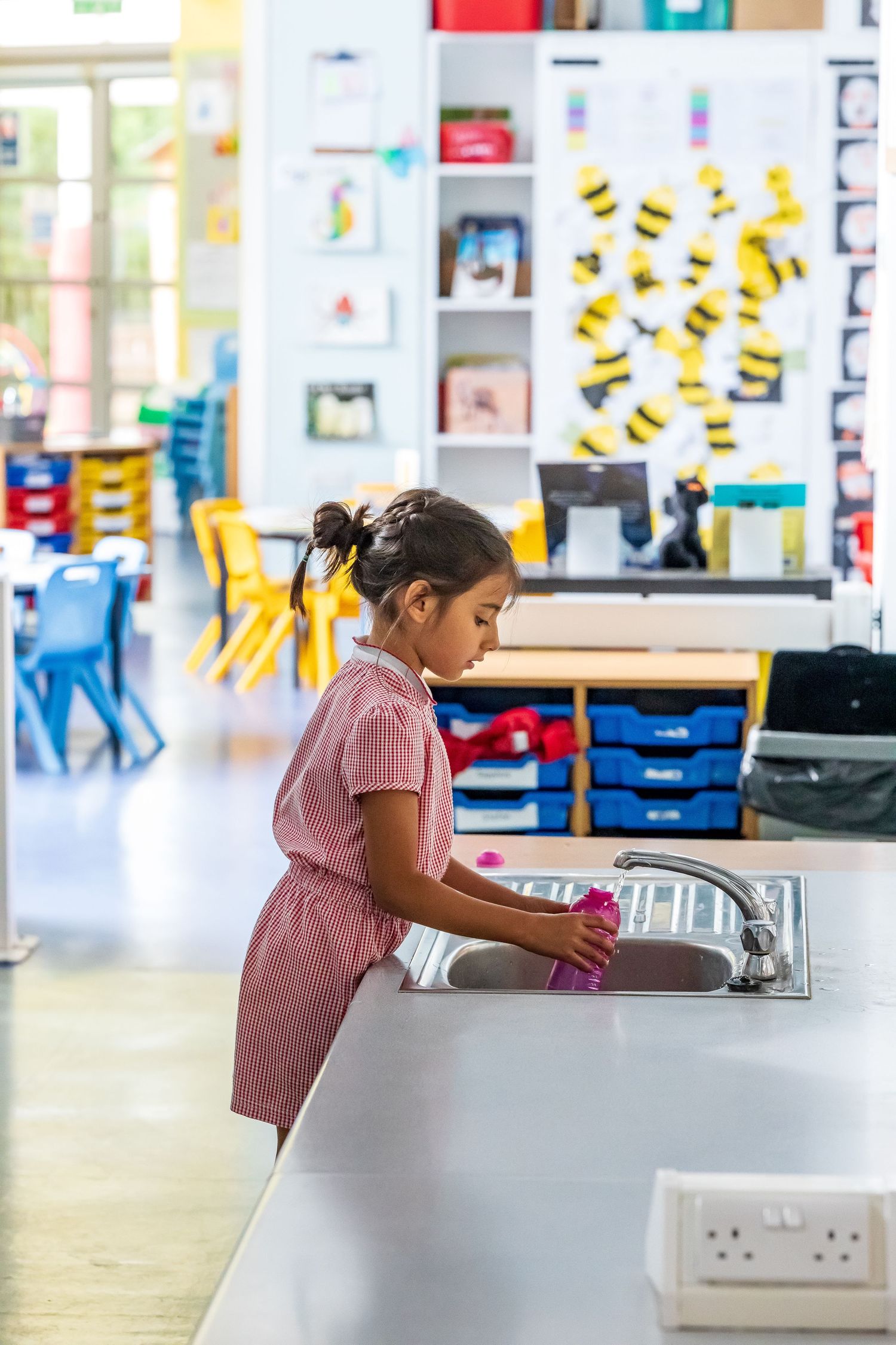 Young girl in pink dress filling water bottle at classroom sink in bright primary school setting.