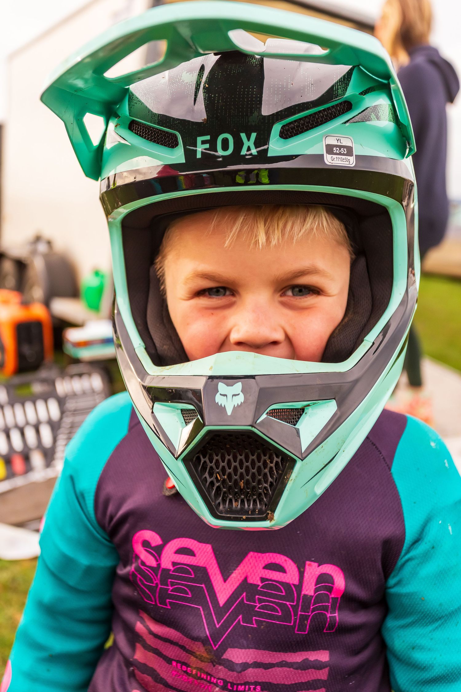 Young child wearing an oversized teal Fox motocross helmet and Seven jersey at an outdoor motorsport event.