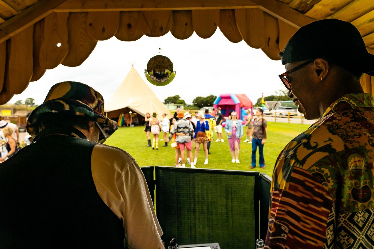 Two DJs performing under a tent canopy overlooking a lively outdoor festival crowd with tipis.