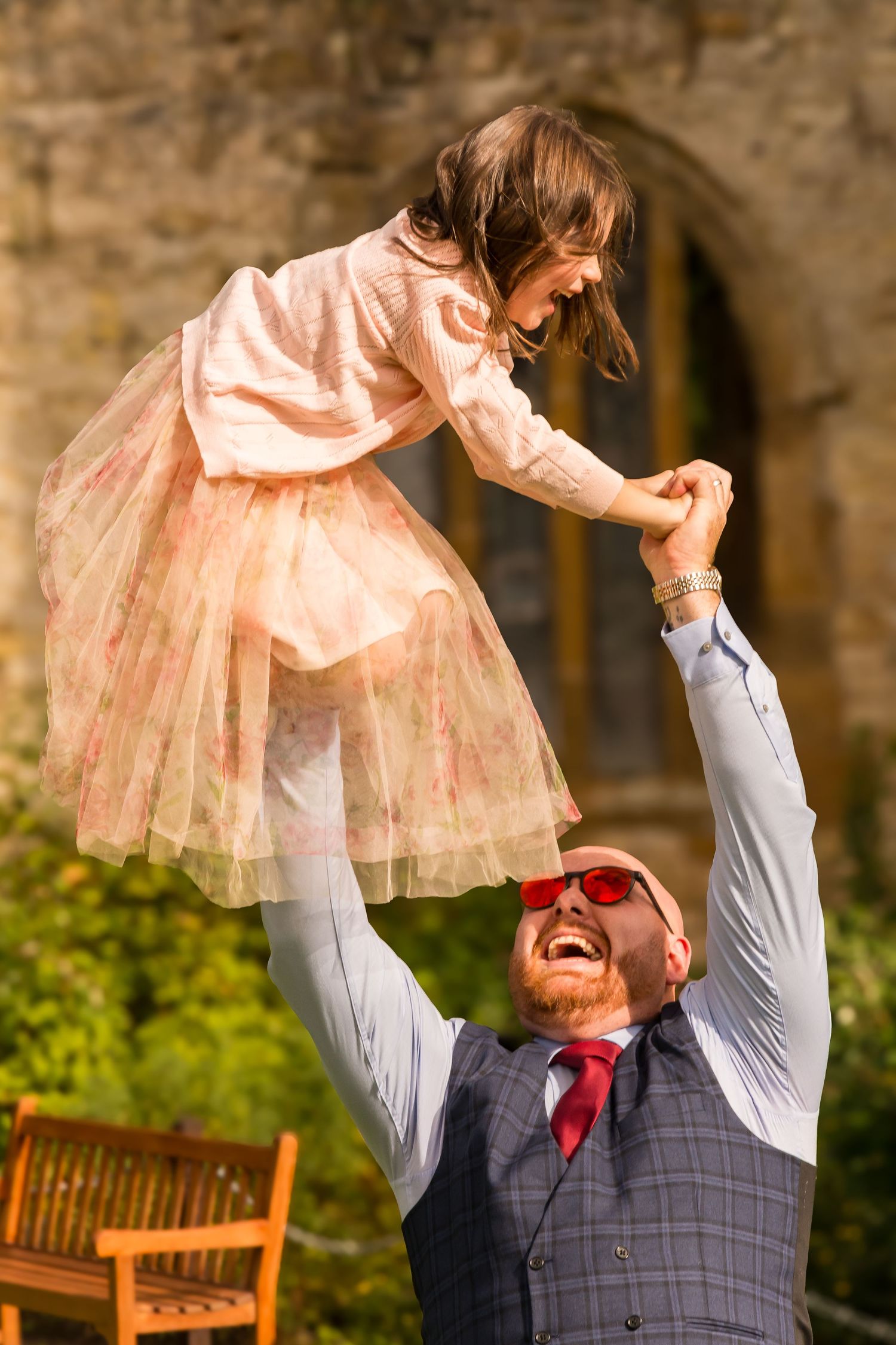 Laughing bearded man in waistcoat lifts joyful girl in pink tulle dress high in the air at a wedding.