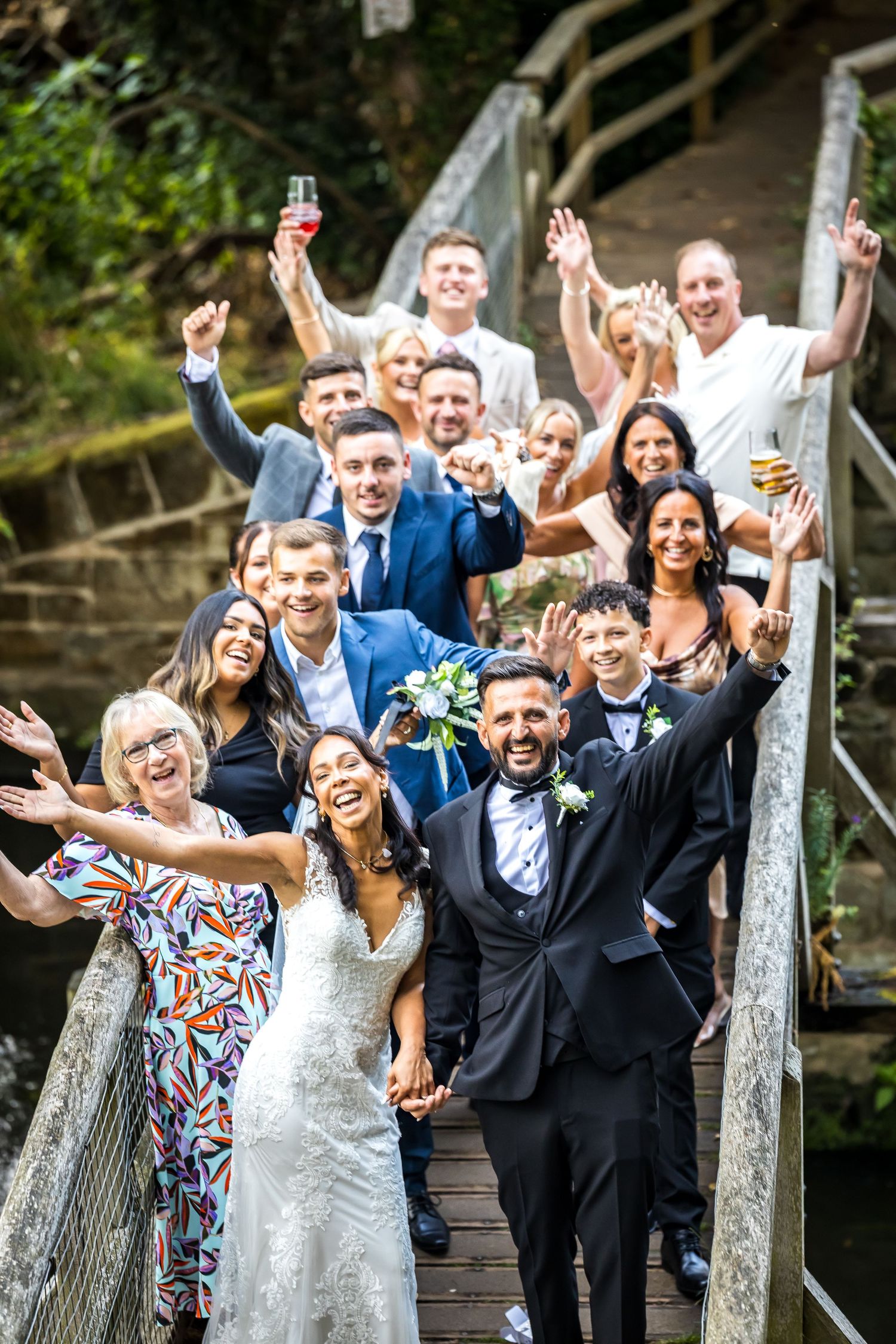 Joyful wedding party group celebrating with raised hands on a rustic wooden bridge.