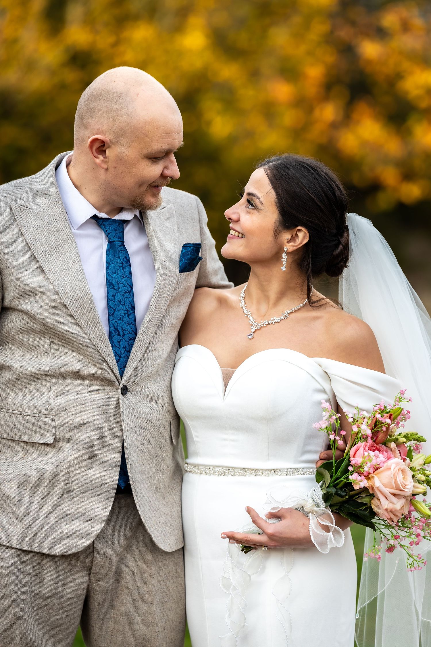 A groom in a grey suit and a bride in a white off-shoulder gown smile at each other in autumn.