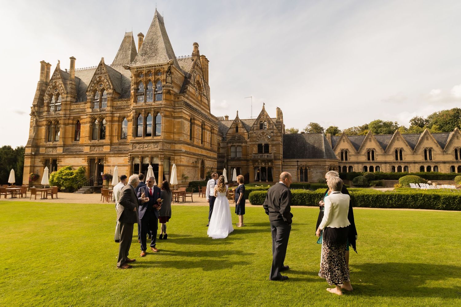 Wedding guests mingle on manicured lawn outside a grand Gothic Victorian manor house on a sunny day.