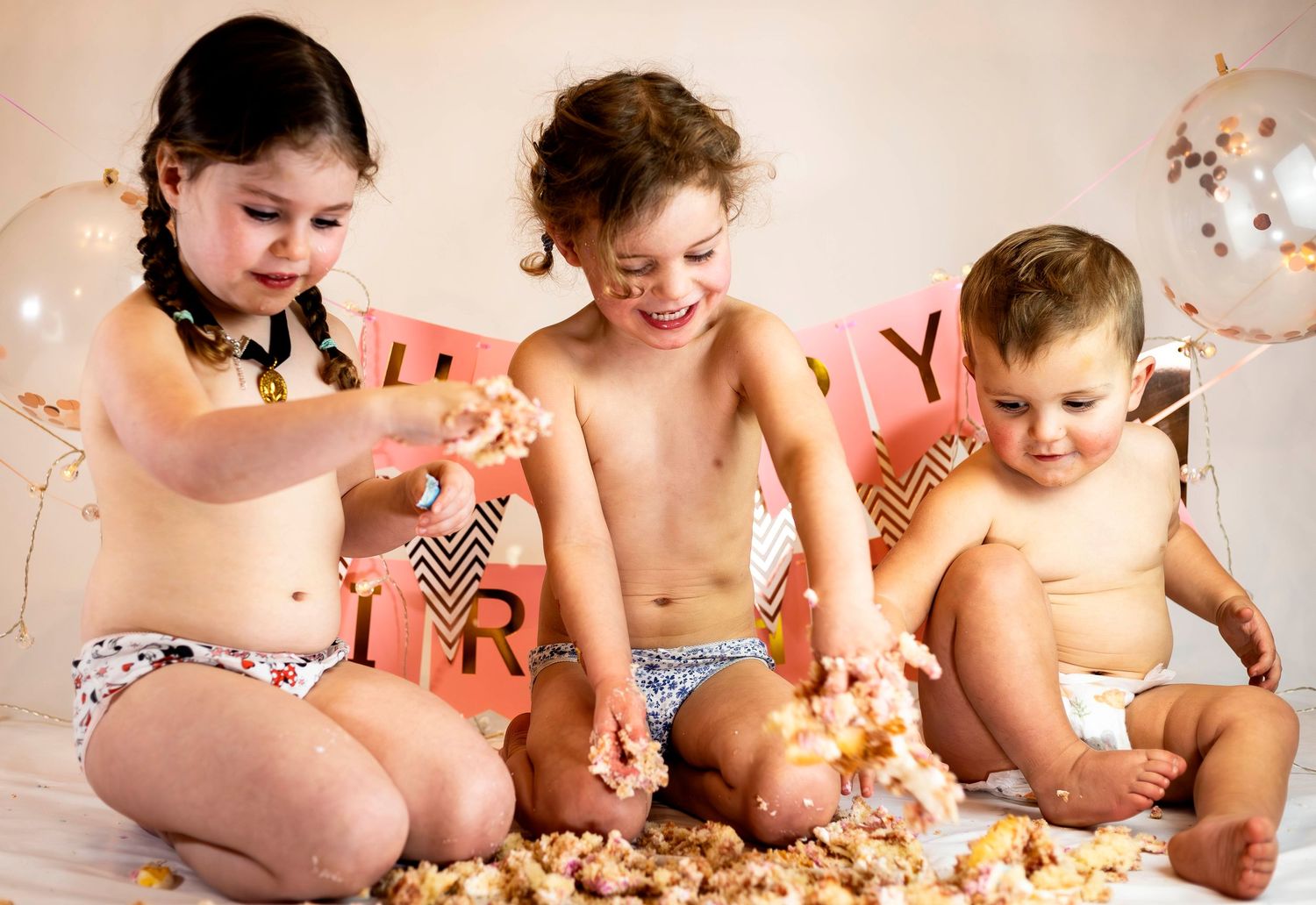 Three young children in diapers joyfully smashing and eating birthday cake together at a party celebration.