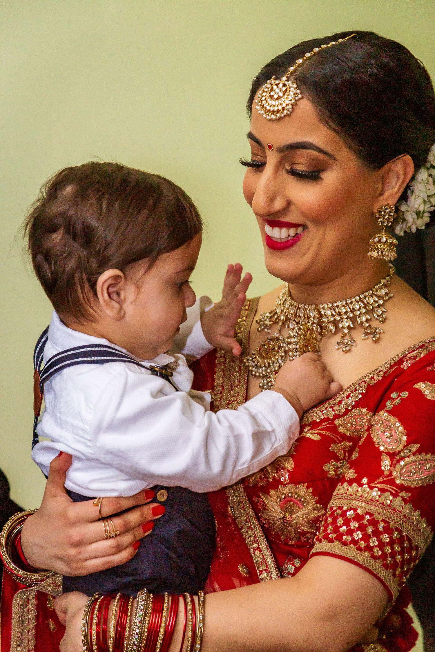 Smiling Indian bride in red lehenga and gold jewelry holding a playful baby boy.