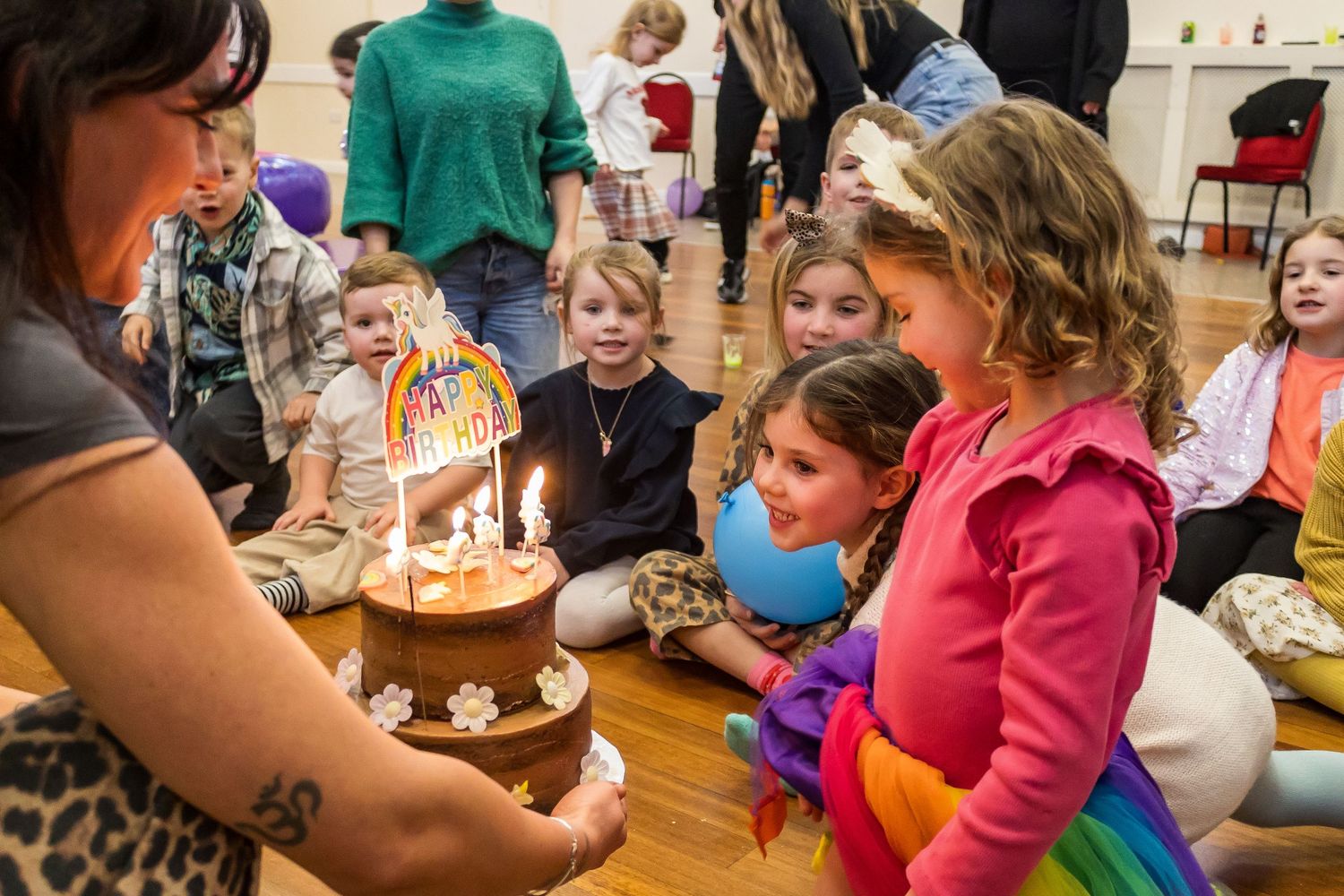 Children gathered around a rainbow birthday cake with lit candles at a kids party.