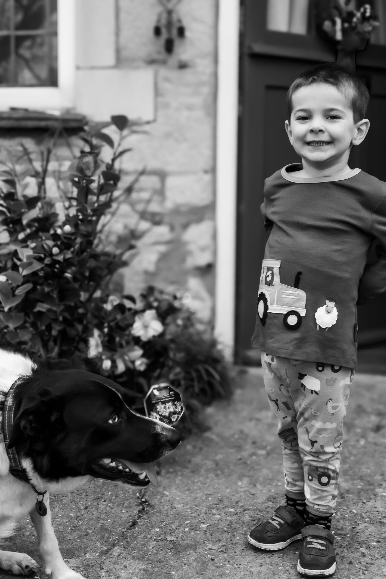 Black and white photo of a happy young boy smiling beside a black and white dog outdoors.