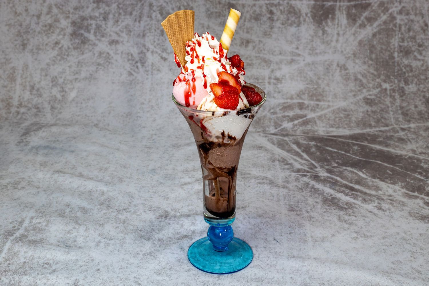 Classic ice cream sundae with chocolate sauce, whipped cream, strawberry topping, and wafer in a blue glass.