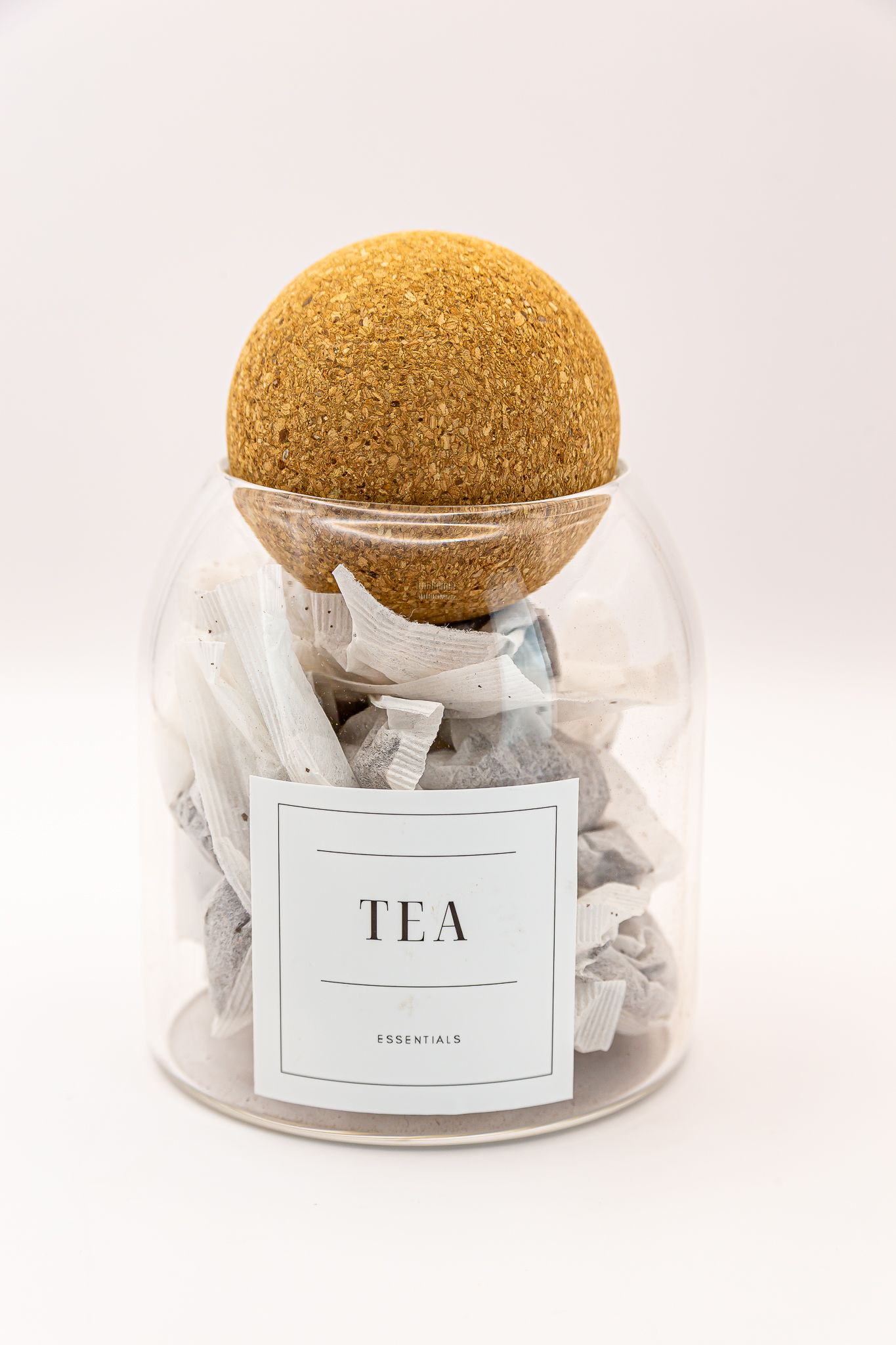 Glass jar labeled TEA filled with tea bags and sealed with a round cork lid on white background. Product photoshoot in studio lighting.