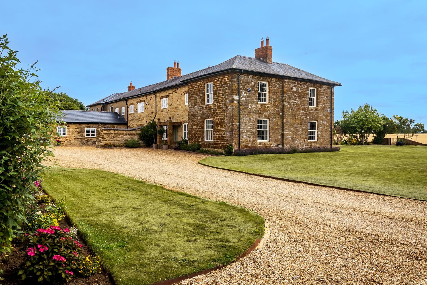 Grand stone country house with manicured lawn, gravel driveway, and colorful flower beds.