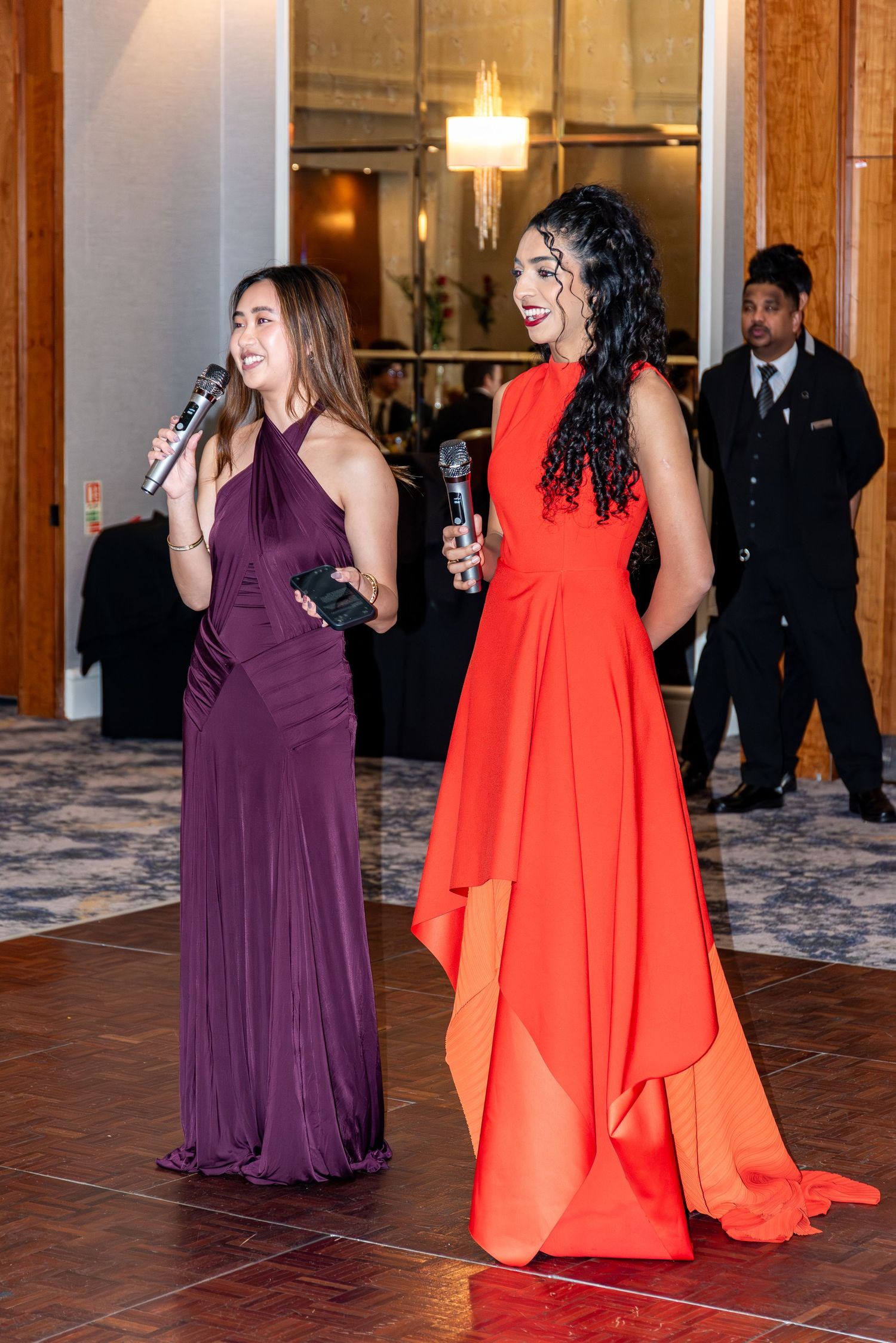 Two elegantly dressed women in purple and red gowns speaking at a school ball.