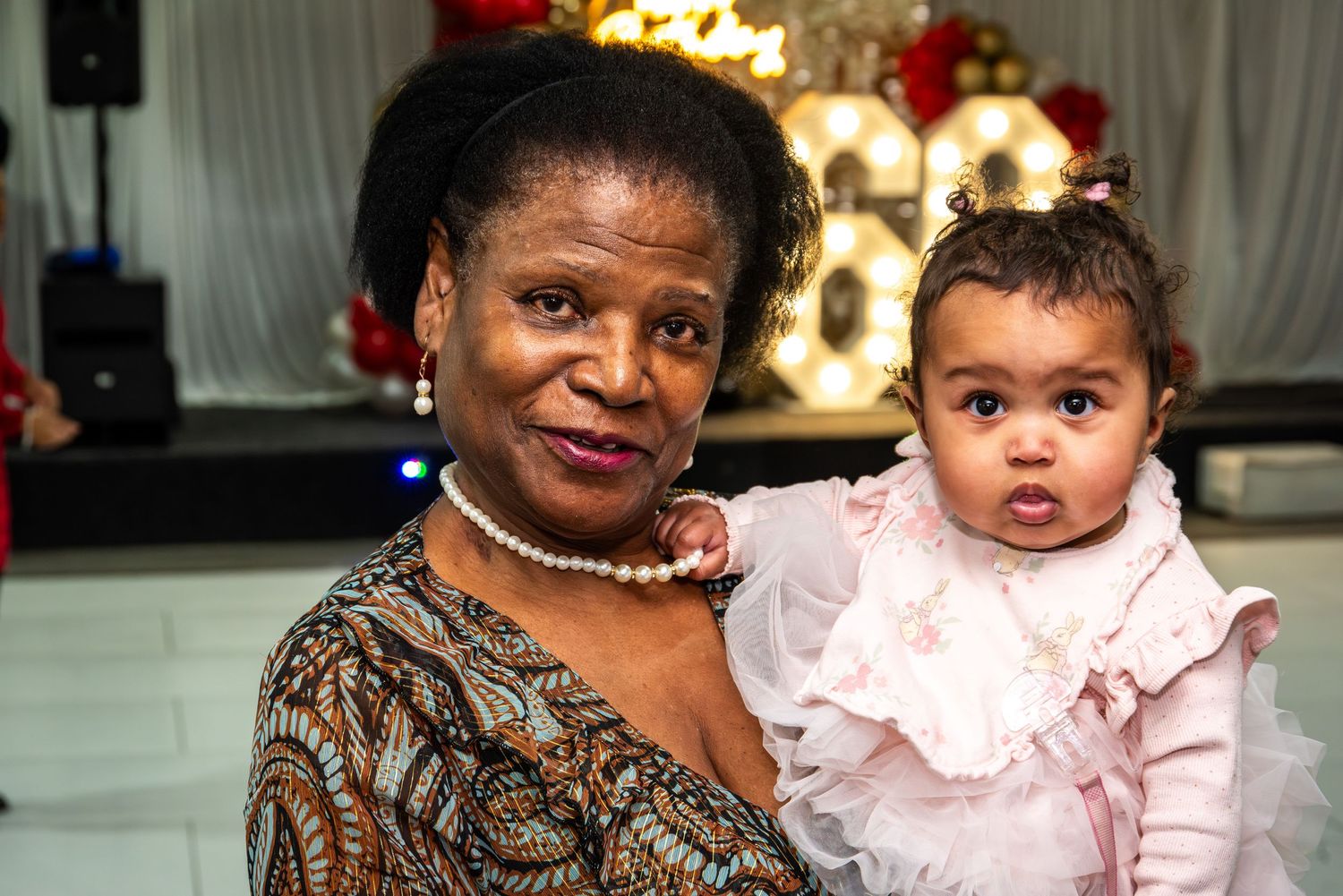 Elderly woman holding a baby girl in a pink dress at a celebration event.