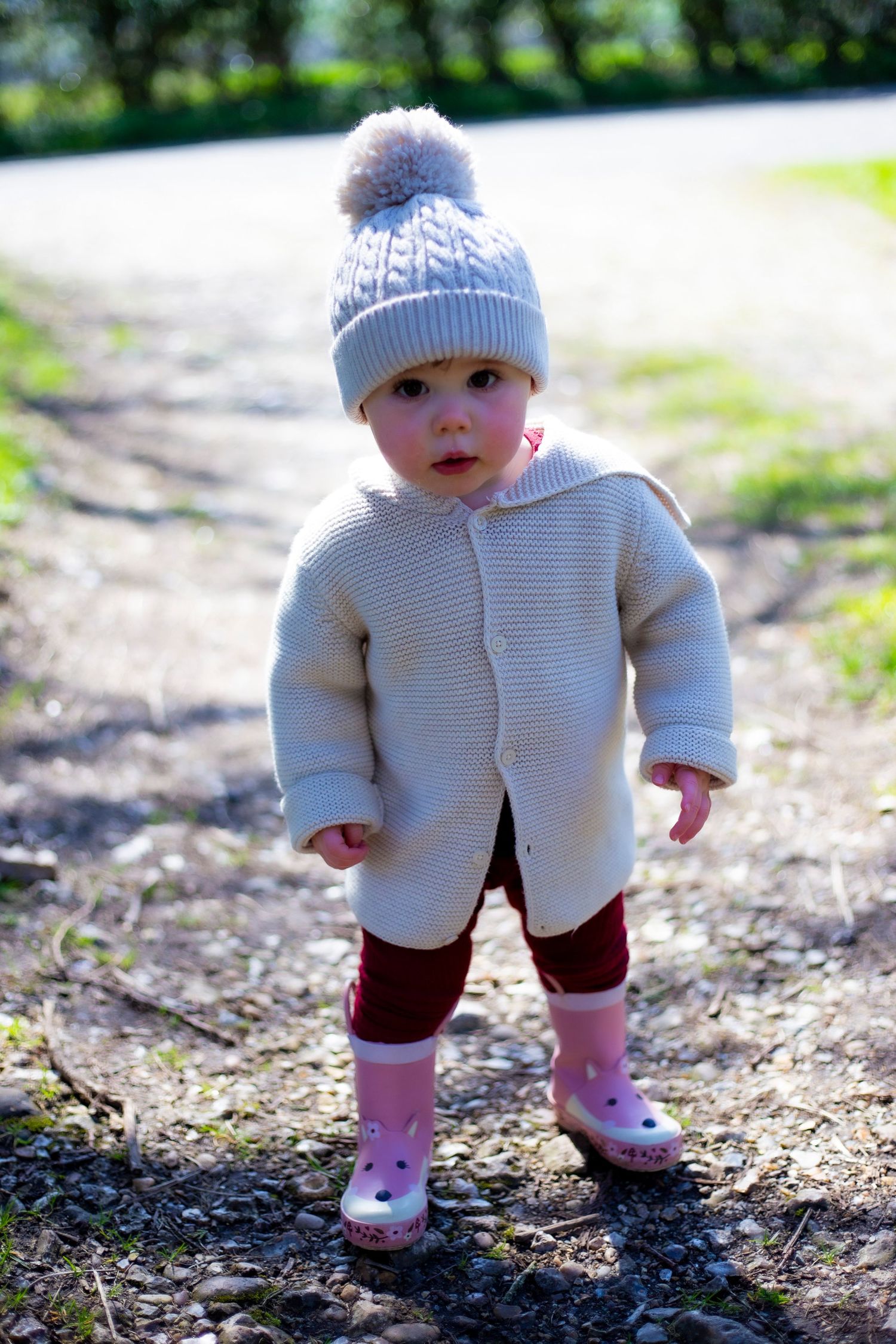 Toddler girl in cream knit sweater, gray pom-pom hat, and pink rain boots standing on a garden path.