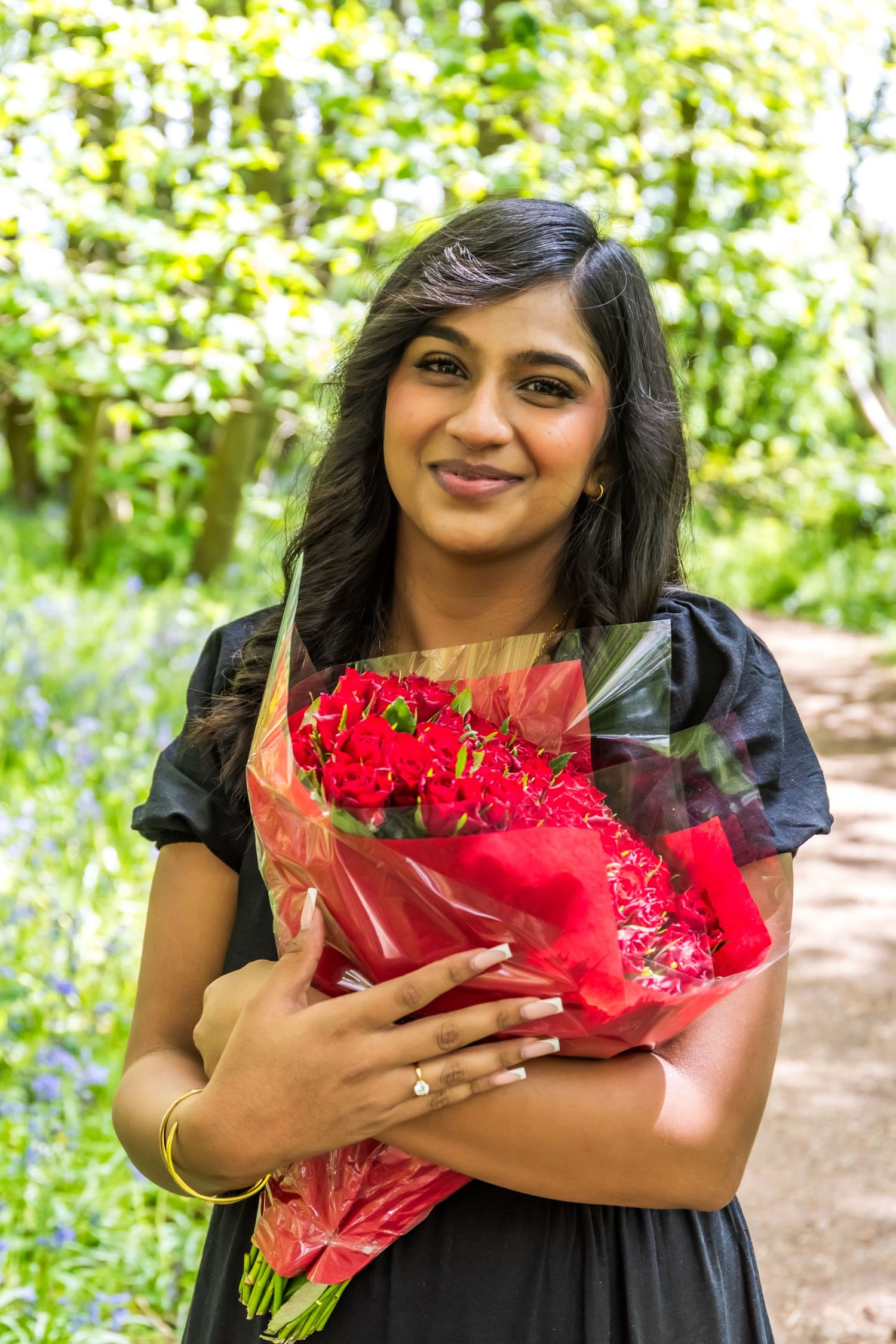 Young woman in black dress holding a bouquet of red roses, smiling outdoors in a green forest setting.