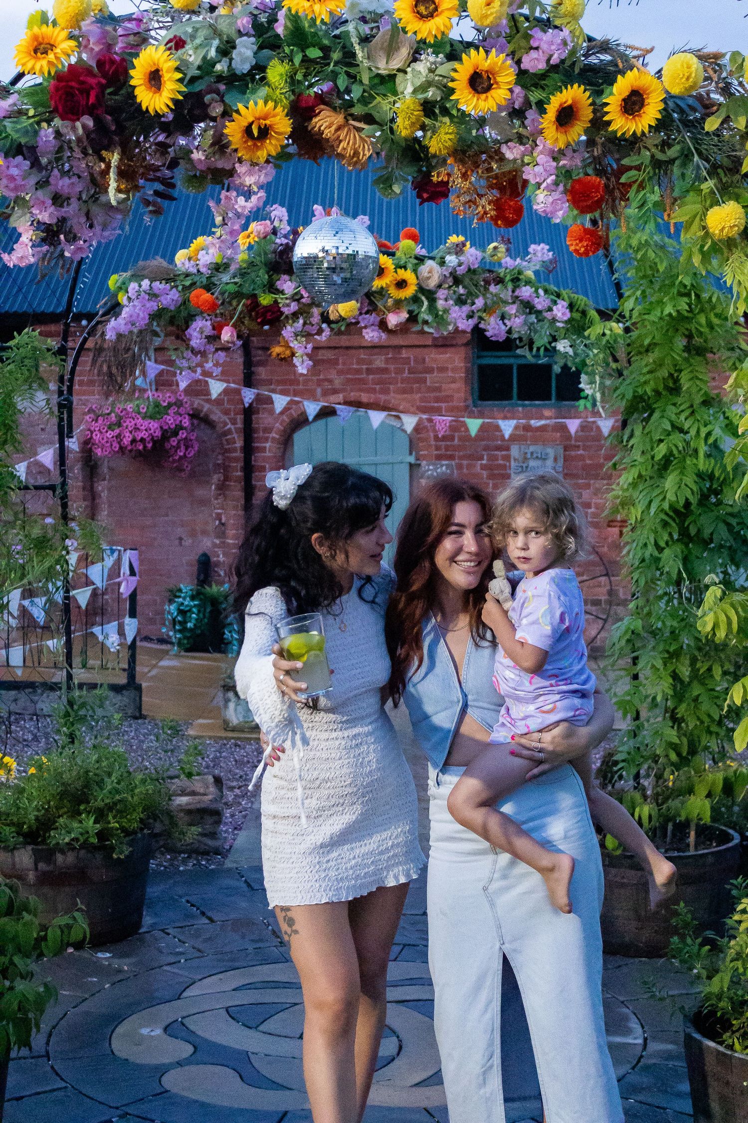 Two women and a young child posing under a colorful floral arch with a disco ball.