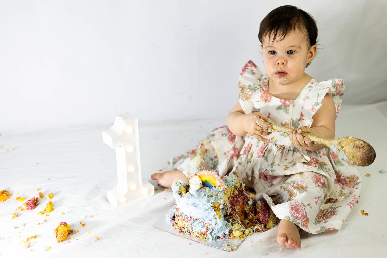 Baby girl in floral dress celebrating first birthday smashing a decorated cake on white background.