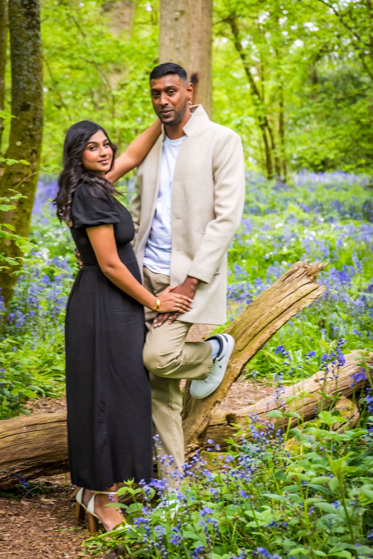 Couple posing in a bluebell woodland, woman in black dress and man in beige jacket leaning on a fallen log.