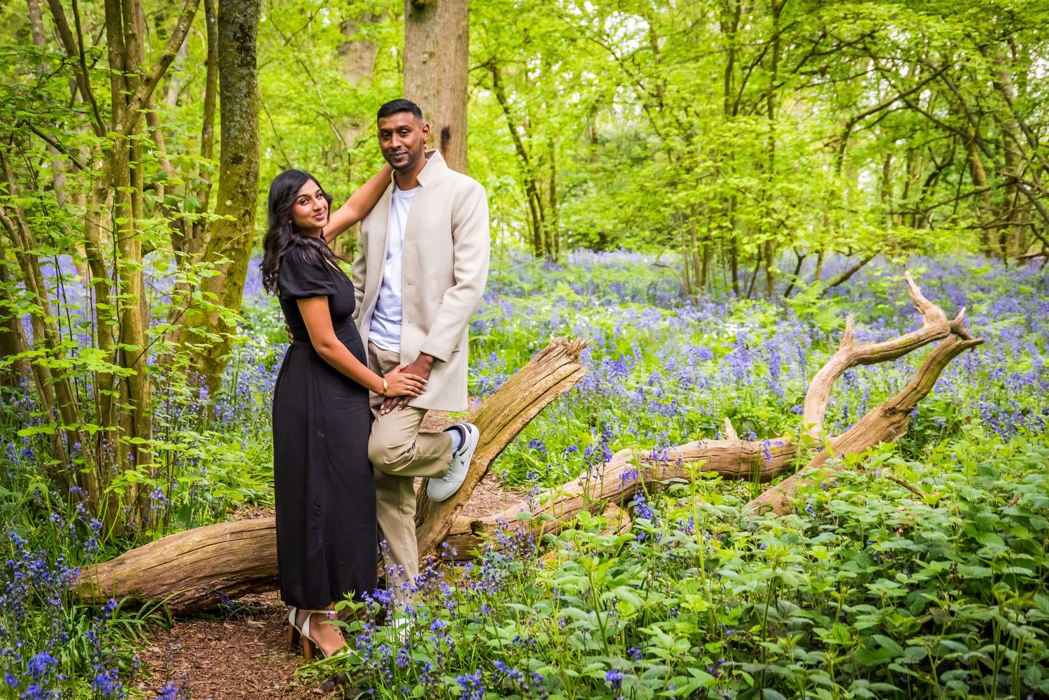 Couple posing romantically in a bluebell woodland, man in cream outfit, woman in black dress, leaning on fallen tree.