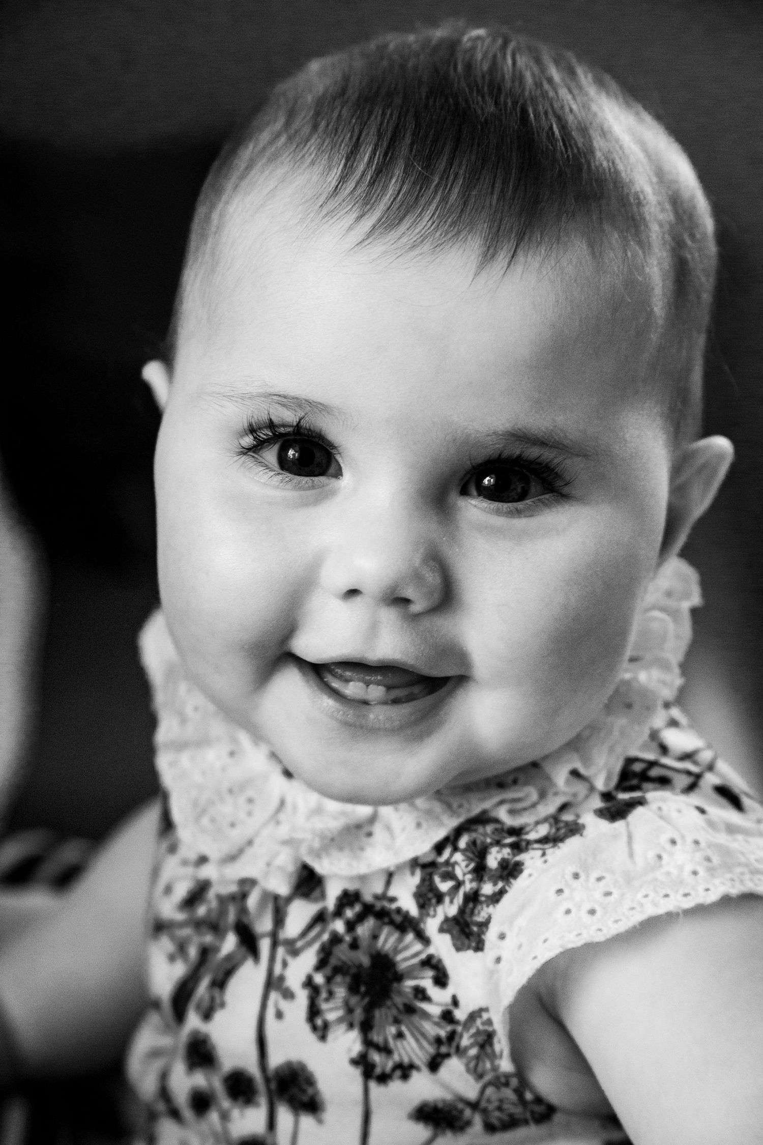 A smiling baby girl in a floral dress poses for a black and white portrait photo.