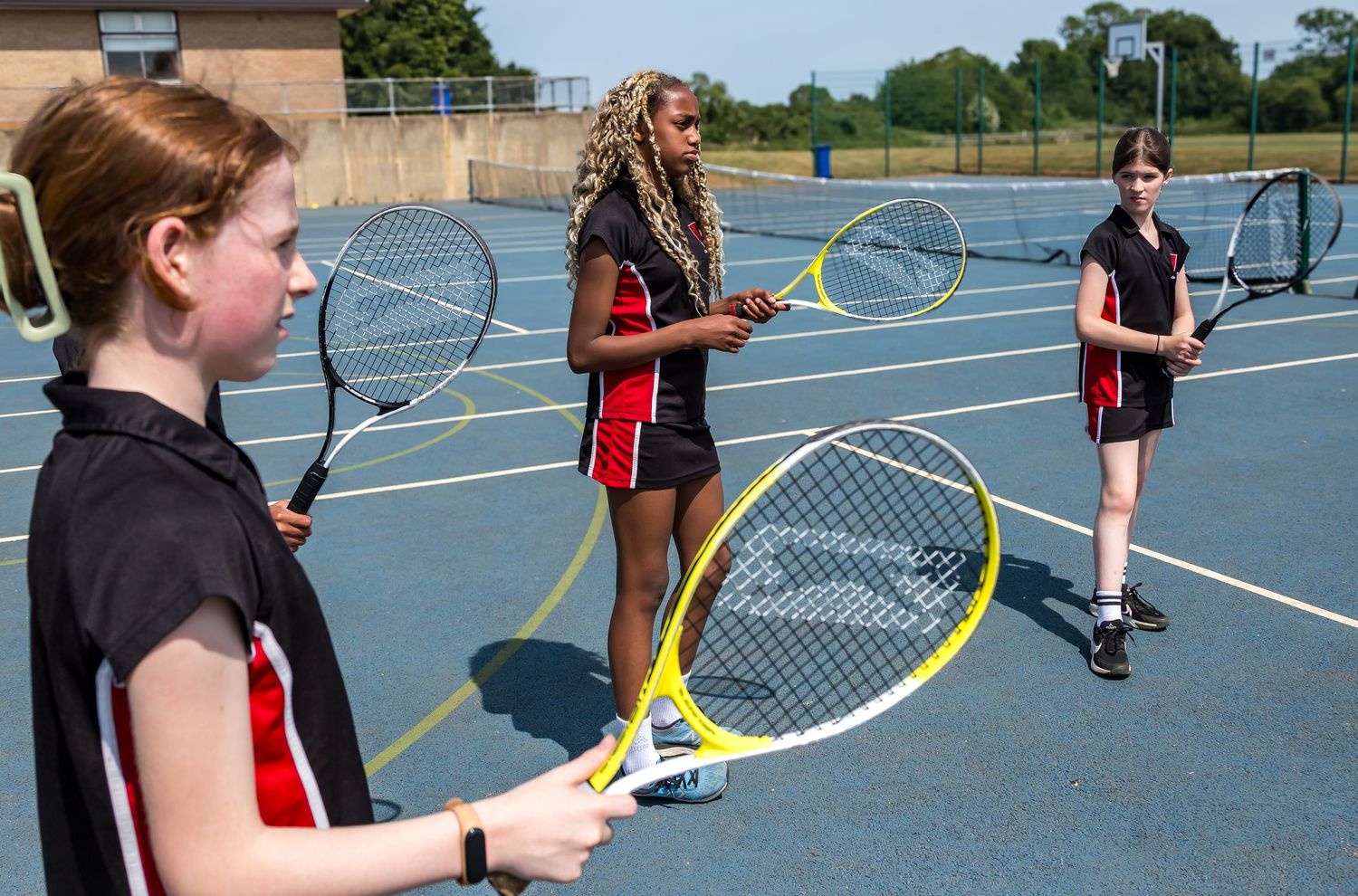 Three girls practicing tennis on a blue outdoor court holding rackets on a sunny day.