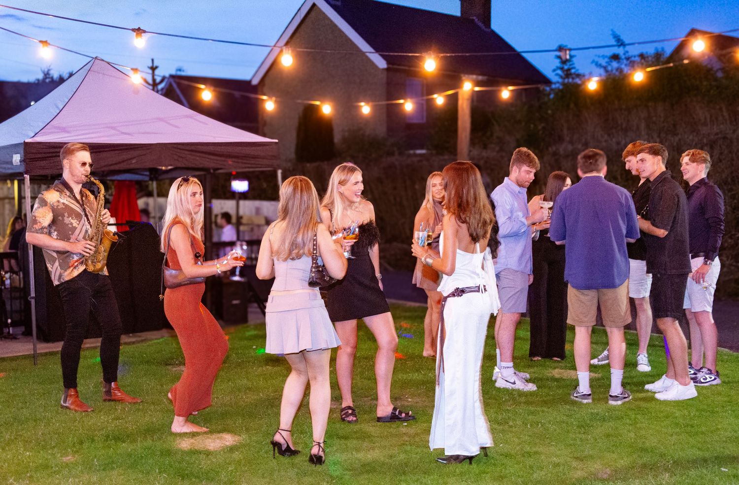 Young adults socializing at an outdoor evening garden party with string lights and a marquee.