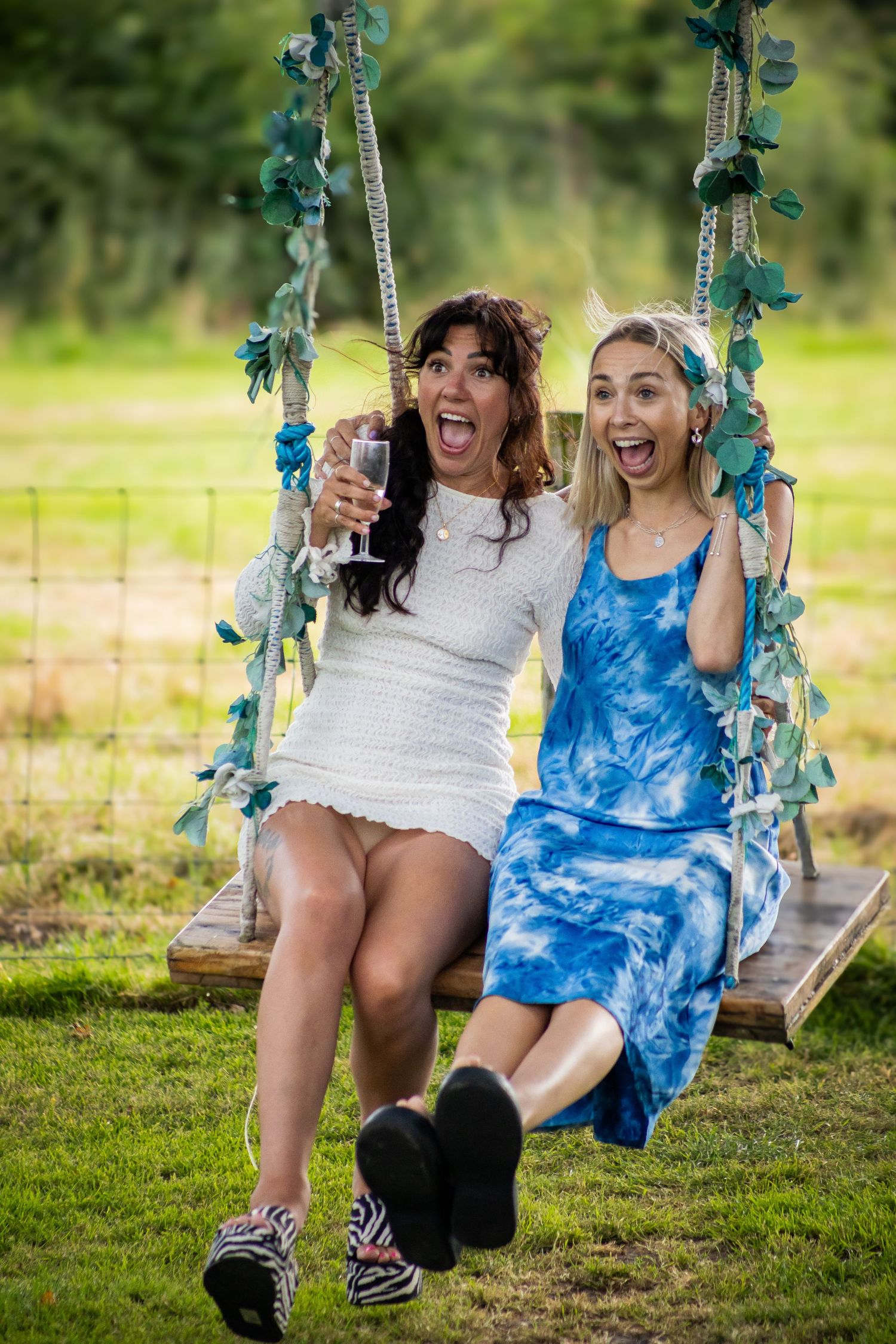 Two women laughing and sitting together on a flower-decorated wooden garden swing holding drinks.