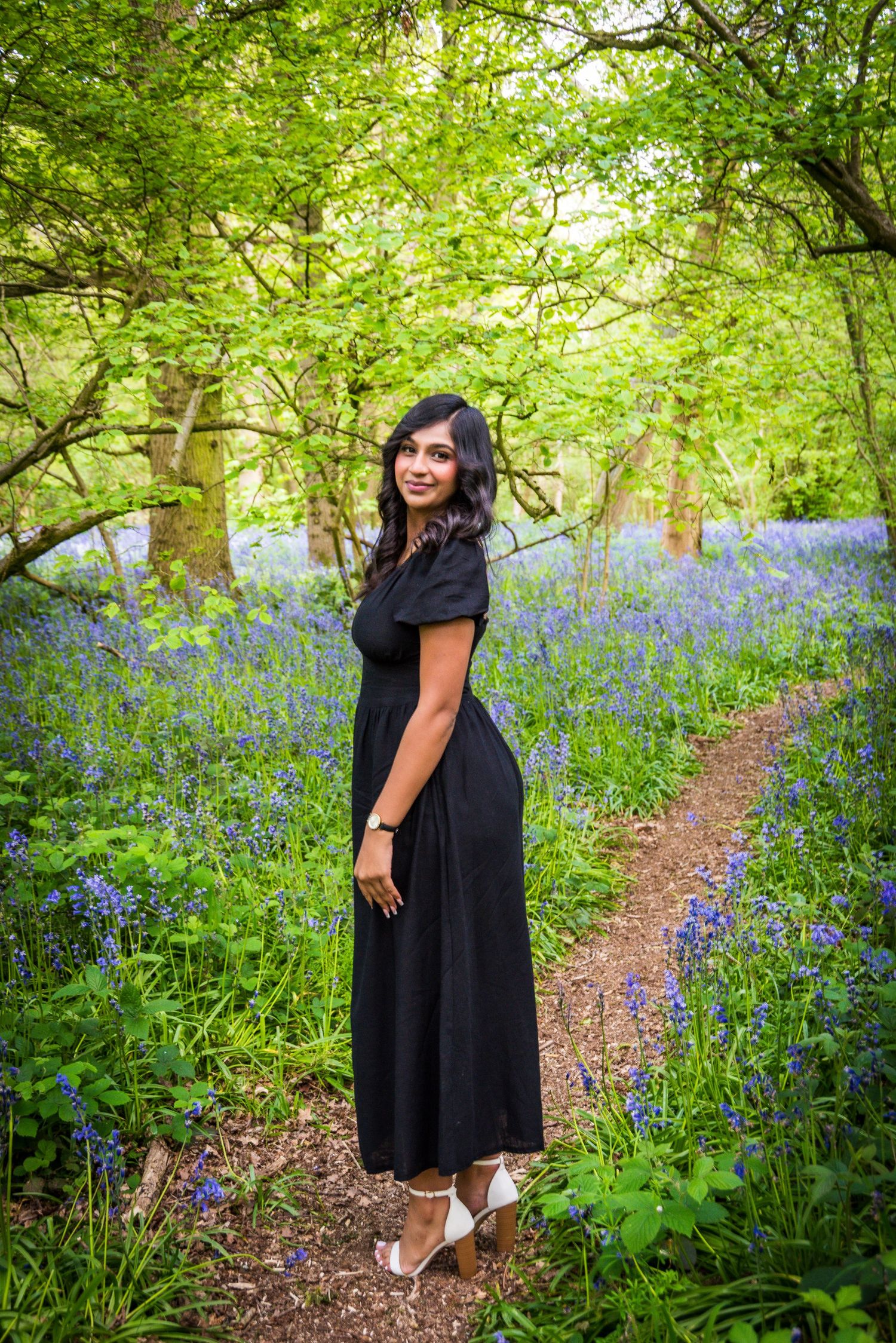 Woman in black midi dress standing on a woodland path surrounded by bluebells.