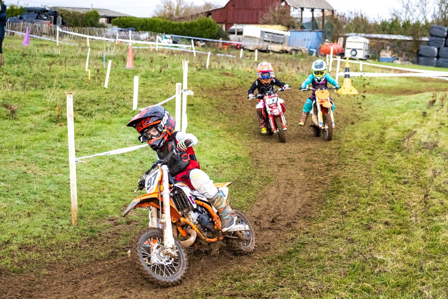 Three young motocross riders race on dirt track on small bikes, wearing colorful helmets and gear on a cloudy day.