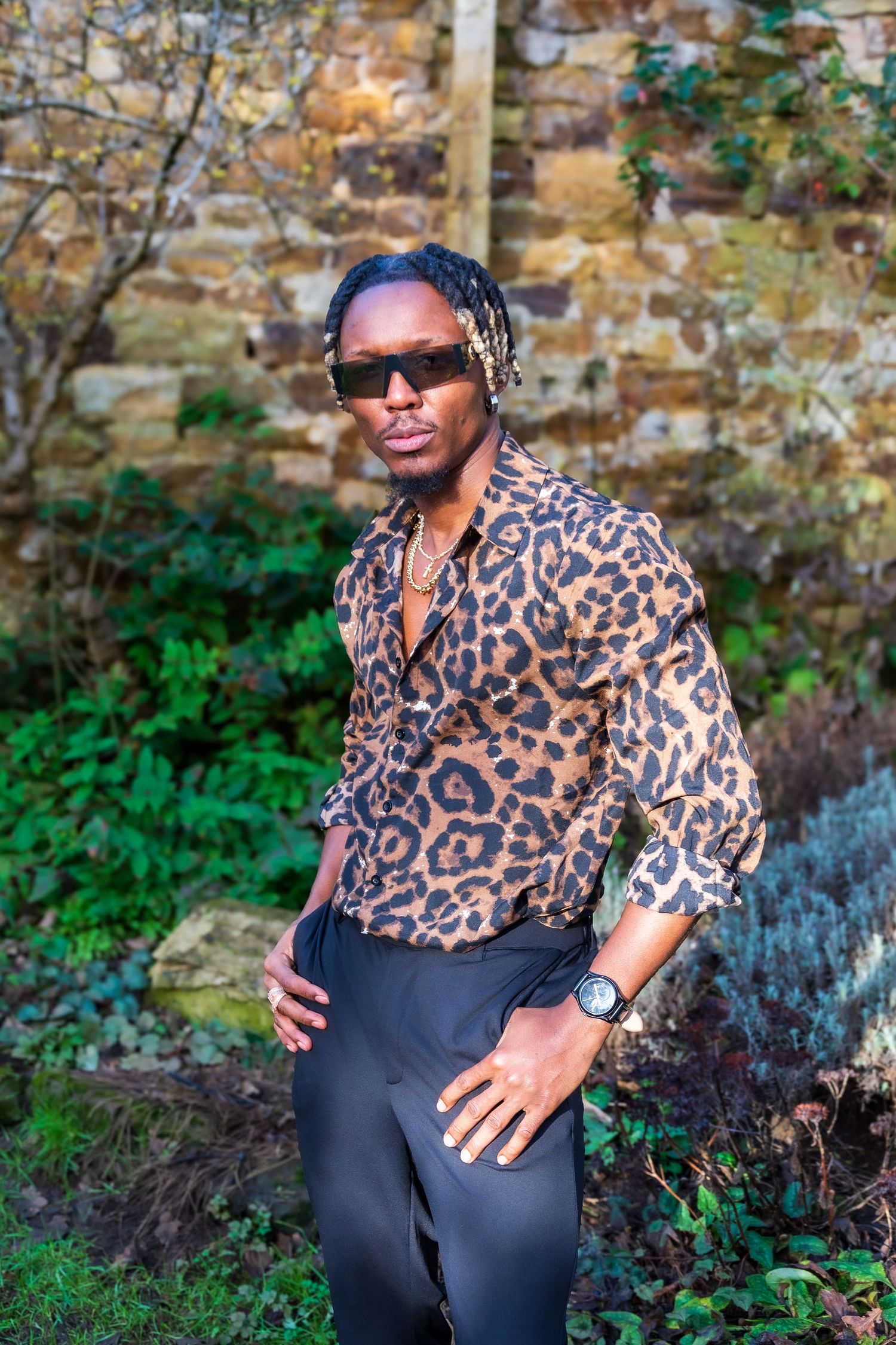 Fashionable man modelling in leopard print shirt and sunglasses posing in a lush garden setting.