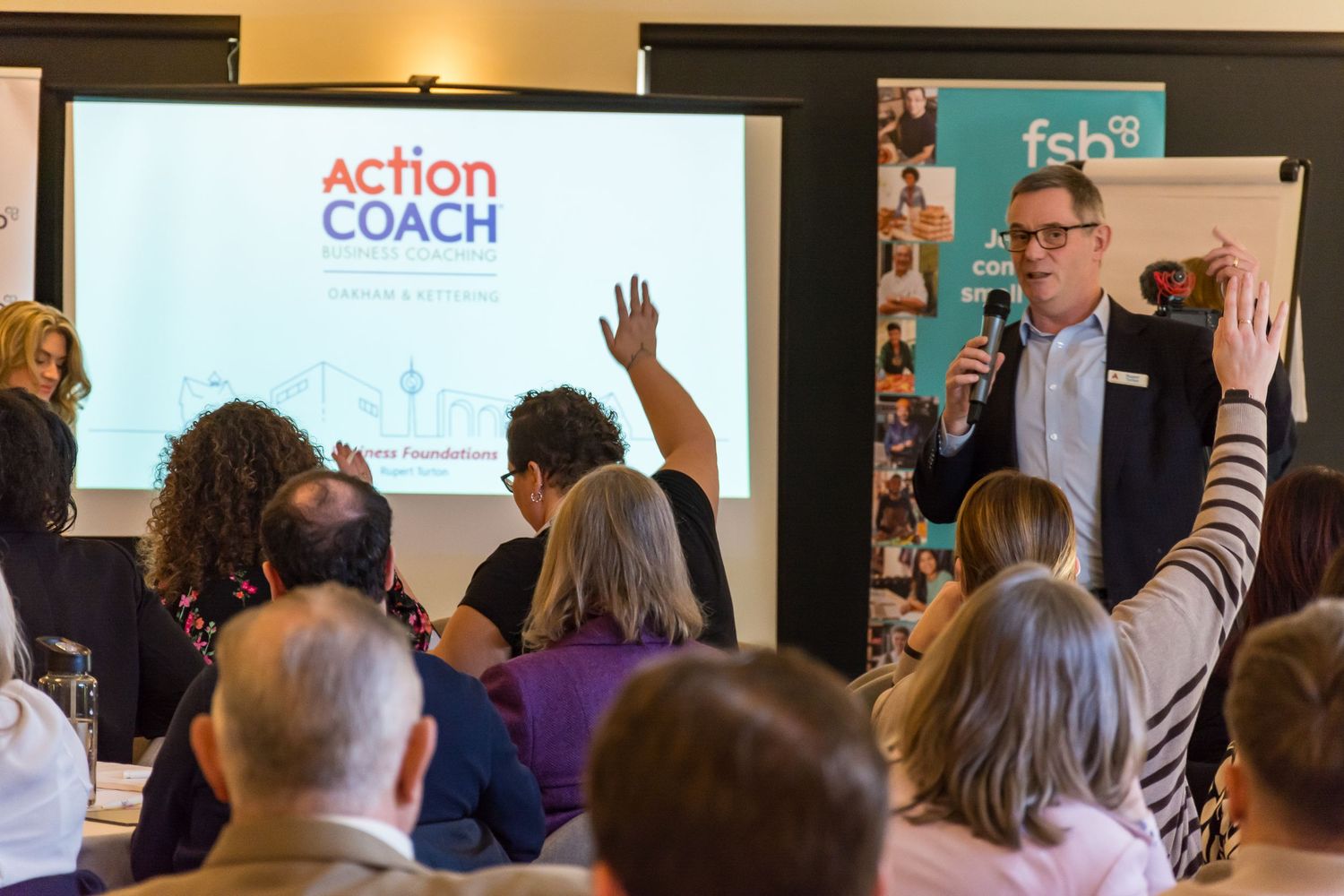 ActionCoach business seminar presenter speaking to engaged audience with raised hands in room.