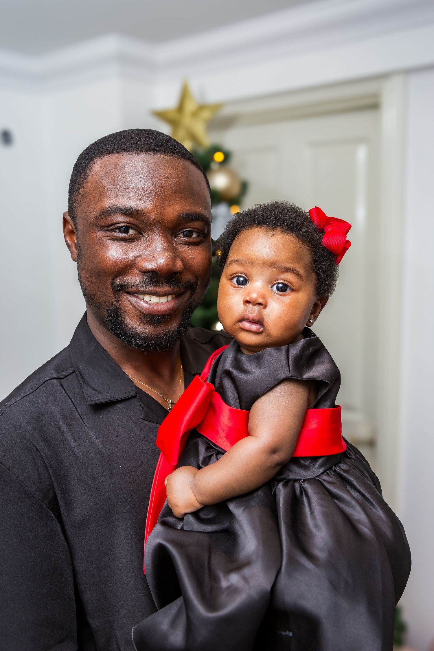 A smiling father holds his baby girl dressed in a black and red bow dress near a Christmas tree.