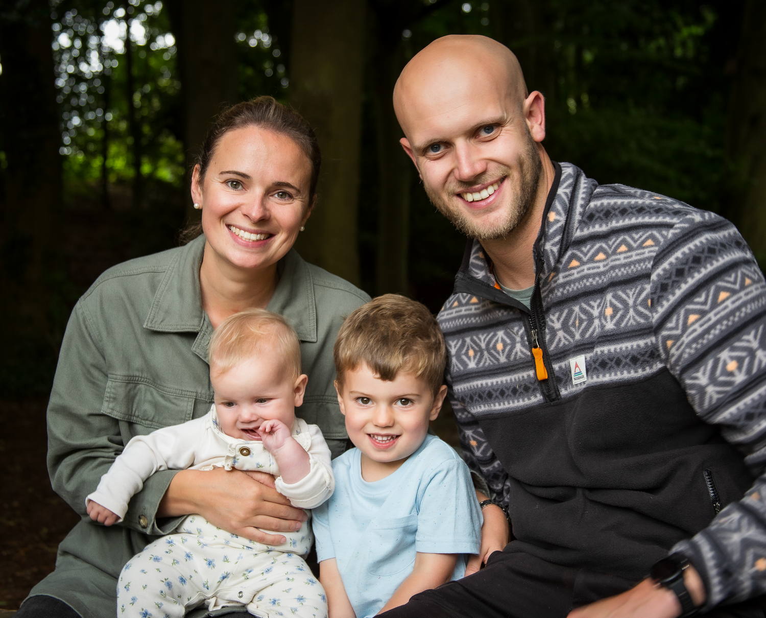 Happy family of four with baby and young boy posing together in a wooded outdoor setting.