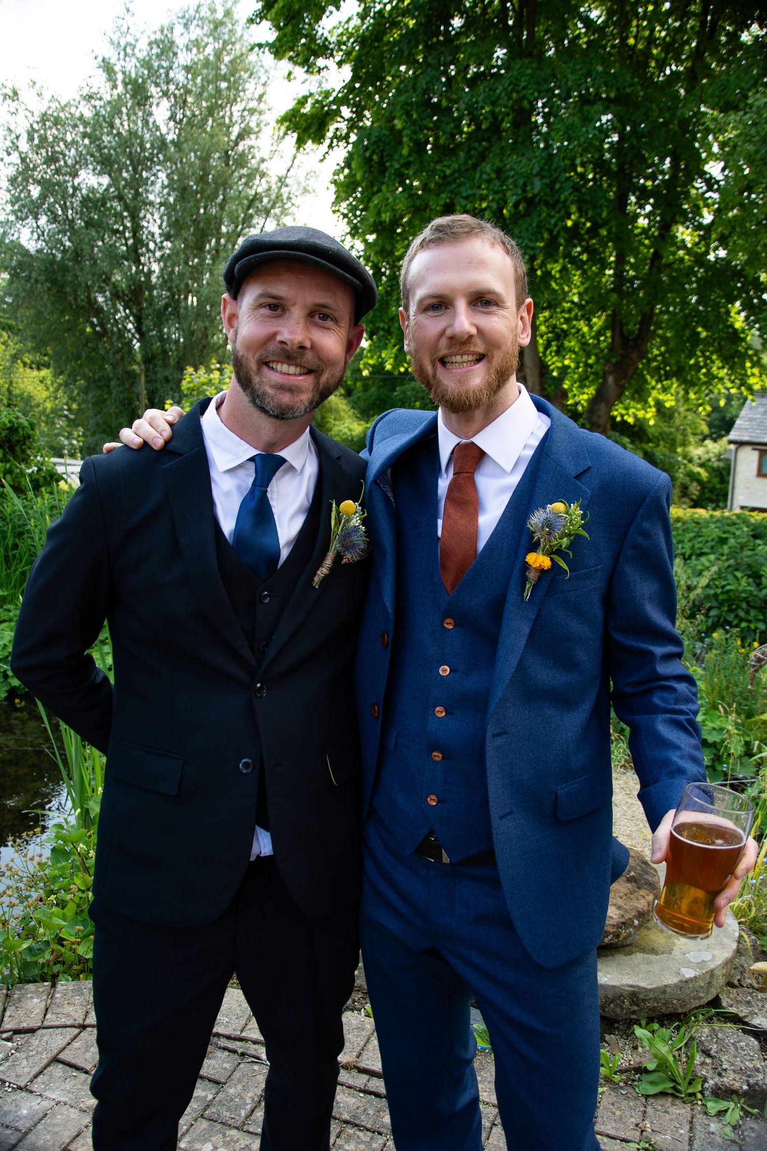 Two smiling men in suits with boutonnieres posing together outdoors at a wedding near a garden pond.
