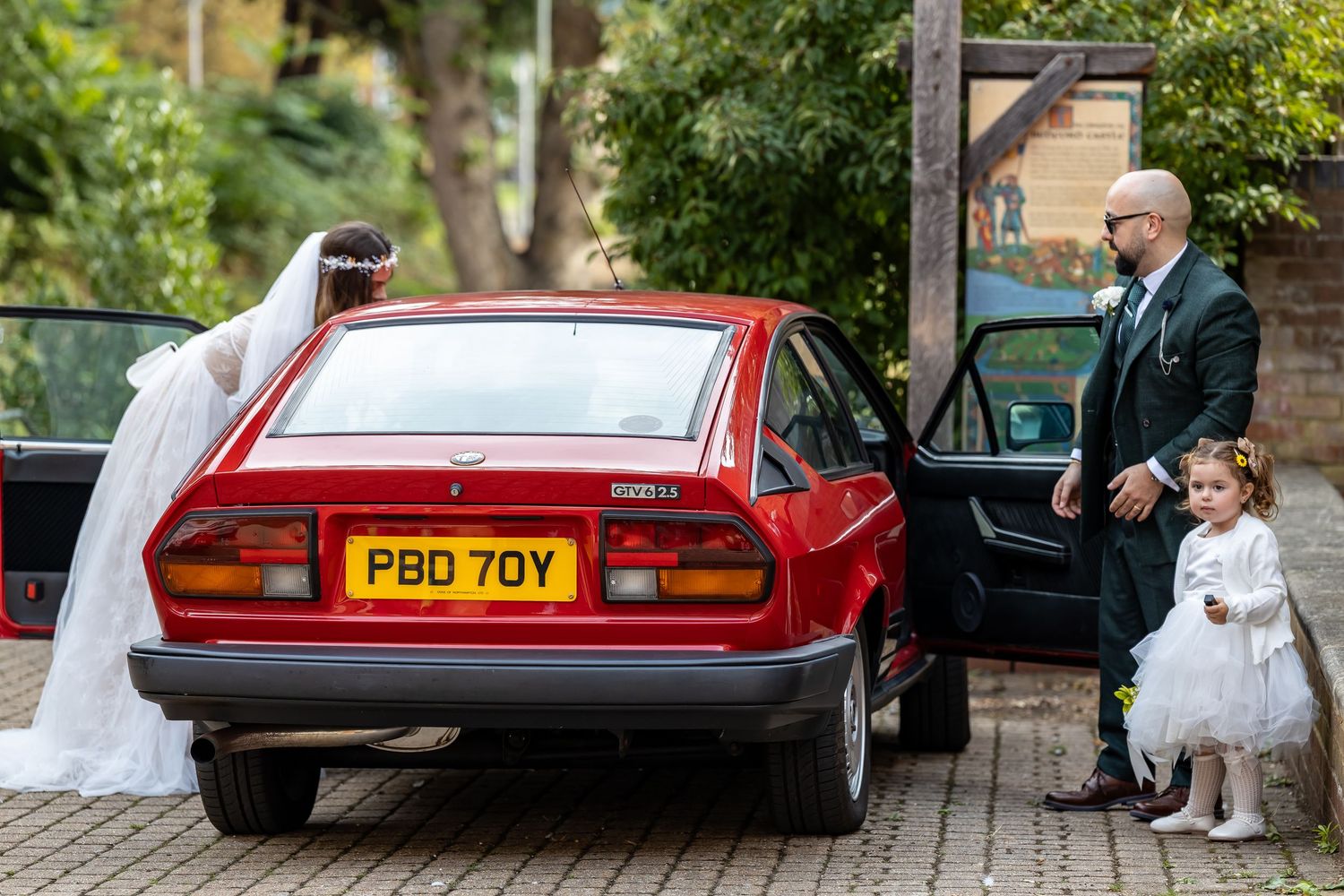 Bride in white gown and flower girl standing beside vintage red car with groom in dark suit at wedding venue.