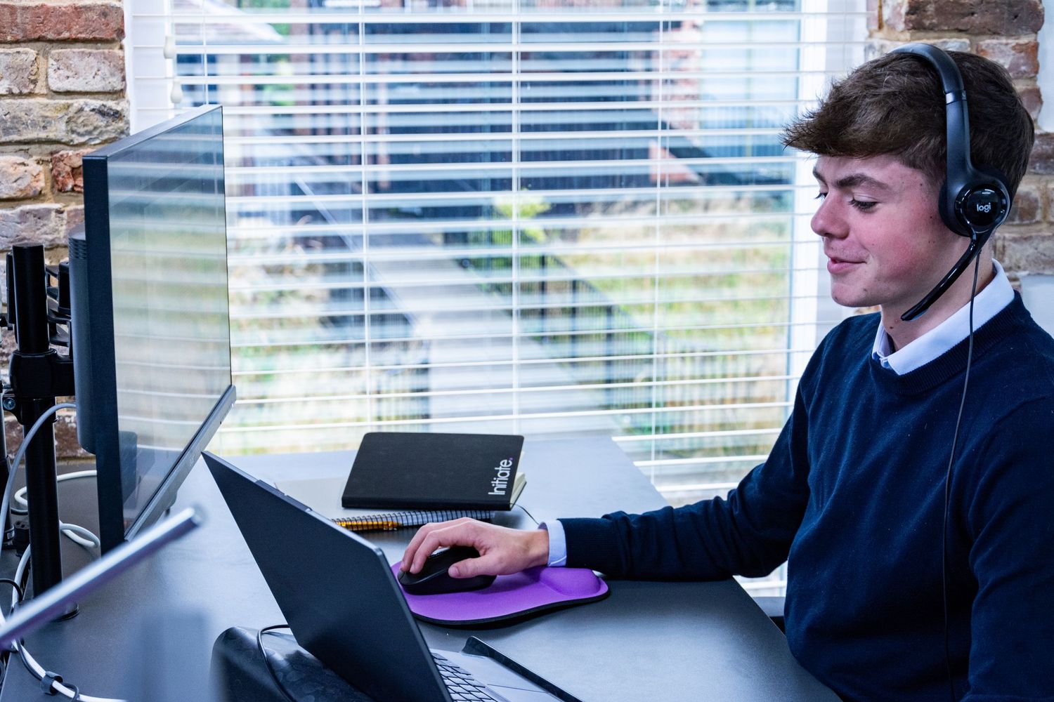 Young male professional wearing headset working on laptop at office desk with dual monitors.