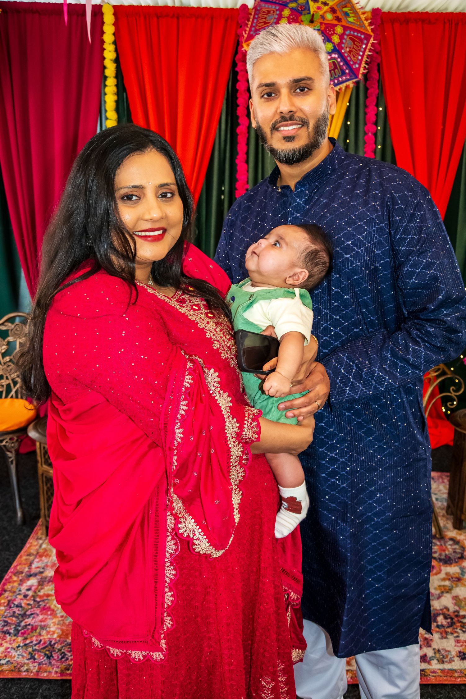 Indian family portrait with parents in traditional attire holding their newborn baby at a festive event.