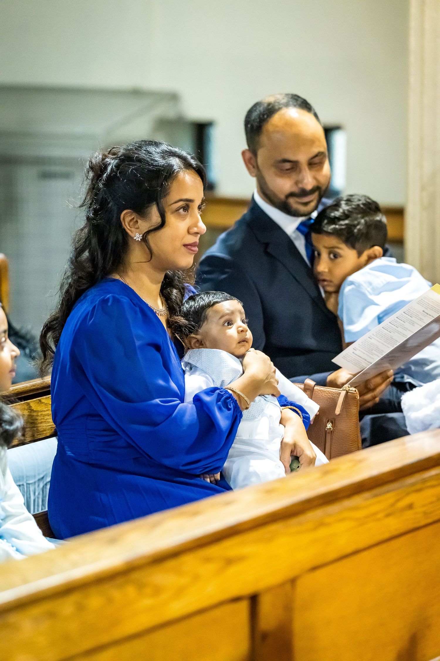 Indian family sitting in church pews during a baptism ceremony, mother holding infant in white outfit.