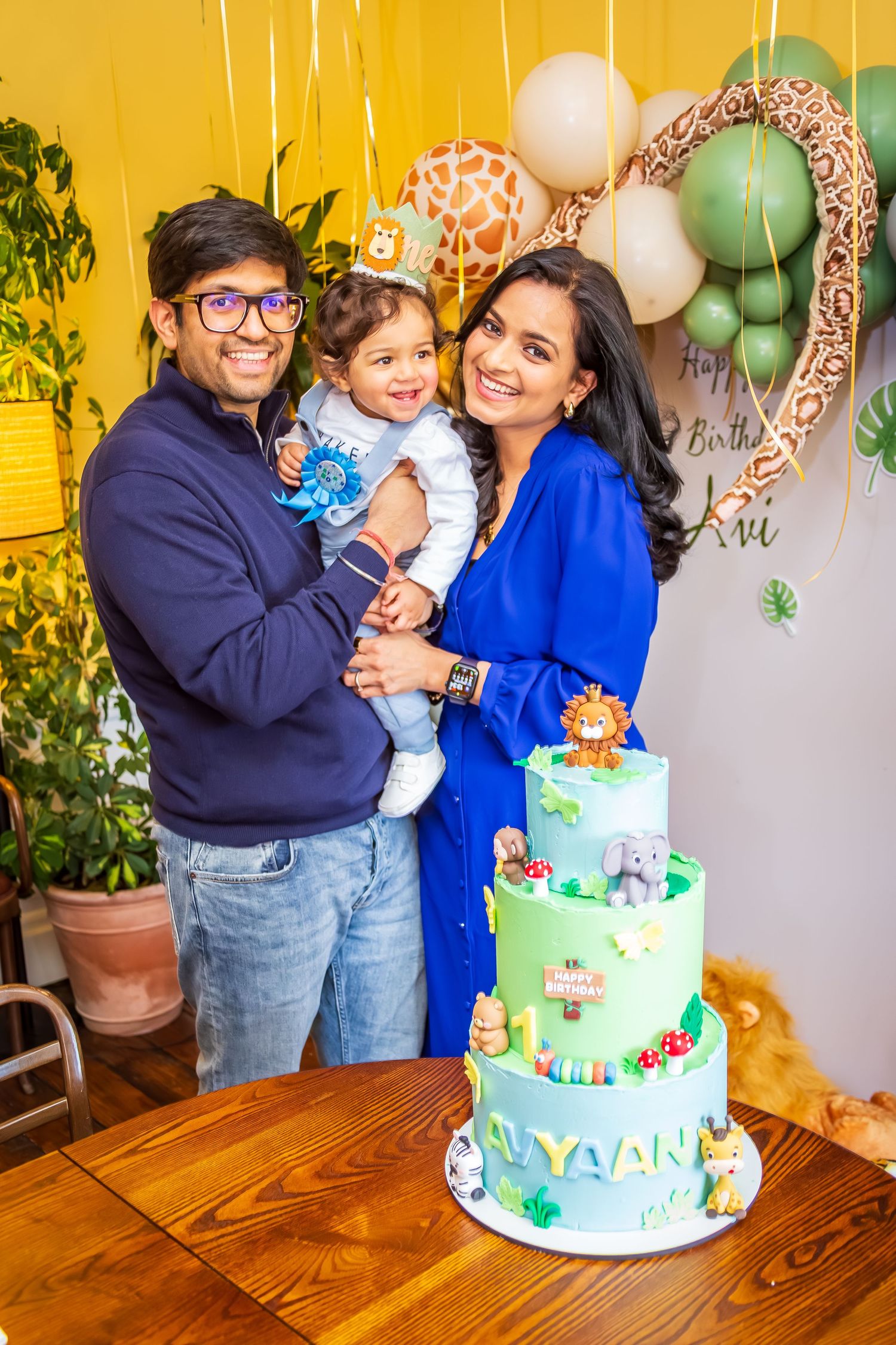 Family of three posing with safari-themed three-tier birthday cake at first birthday celebration.