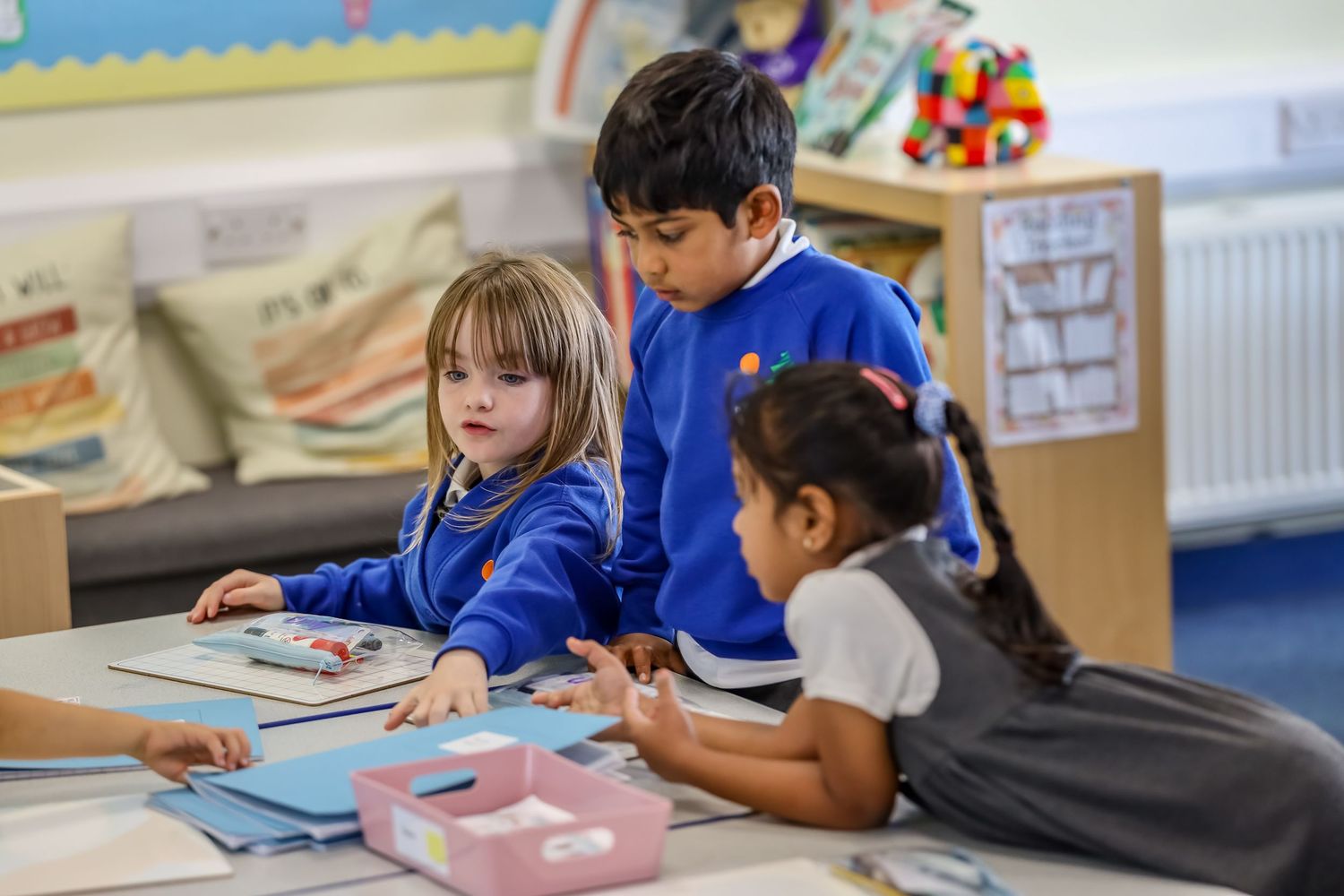 Three primary school children in blue uniforms collaborating on a classroom activity at a table.