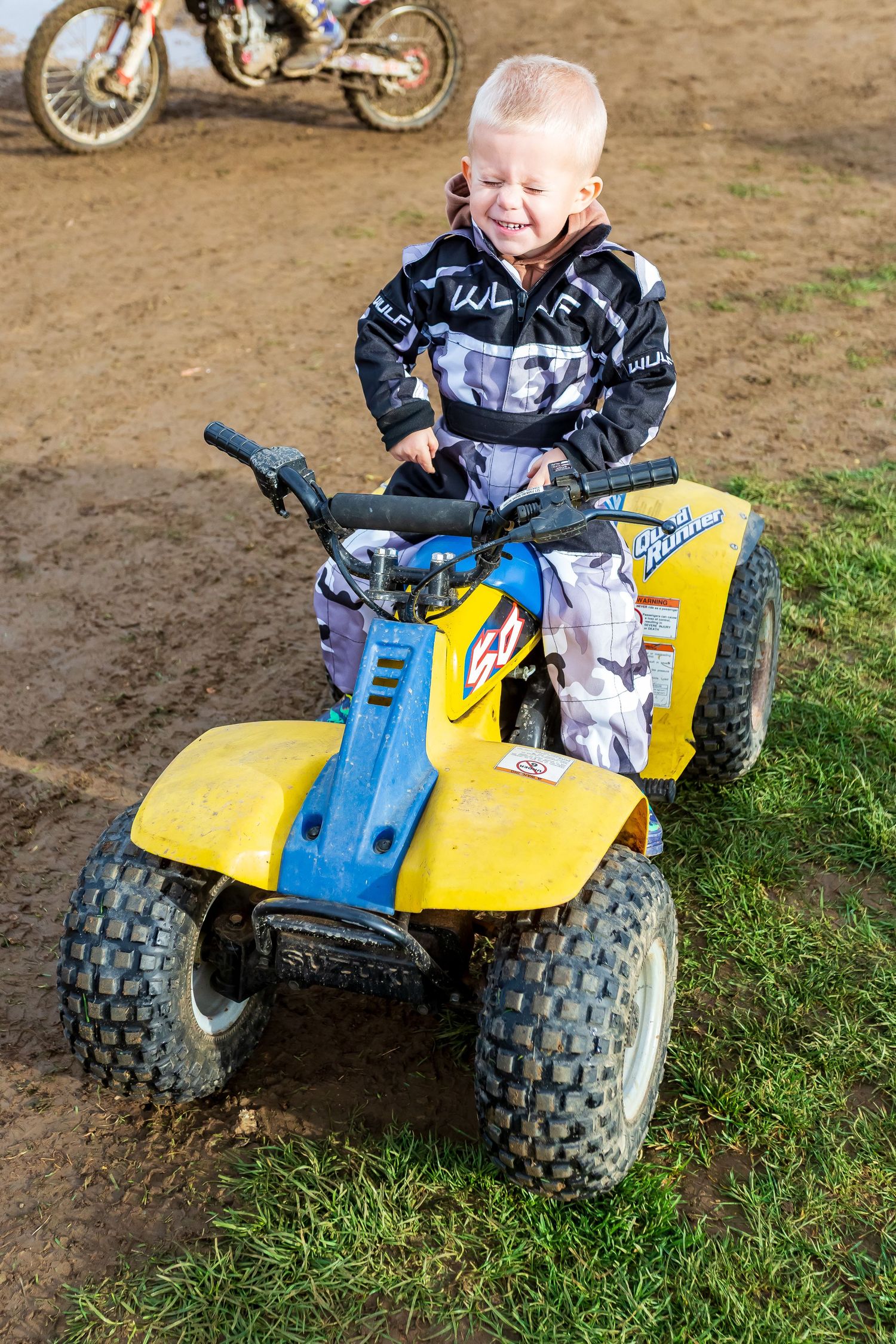 Young boy in motocross gear riding a yellow ATV quad bike on a dirt track with a motorcycle in the background.