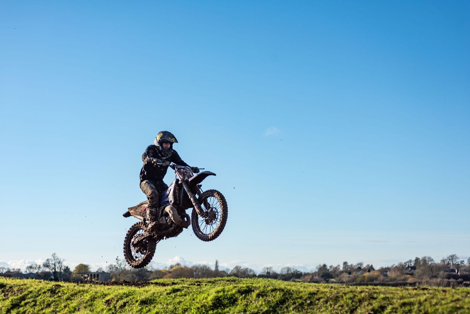 Motocross rider performing a high jump trick on a dirt bike against a clear blue sky on a sunny day.