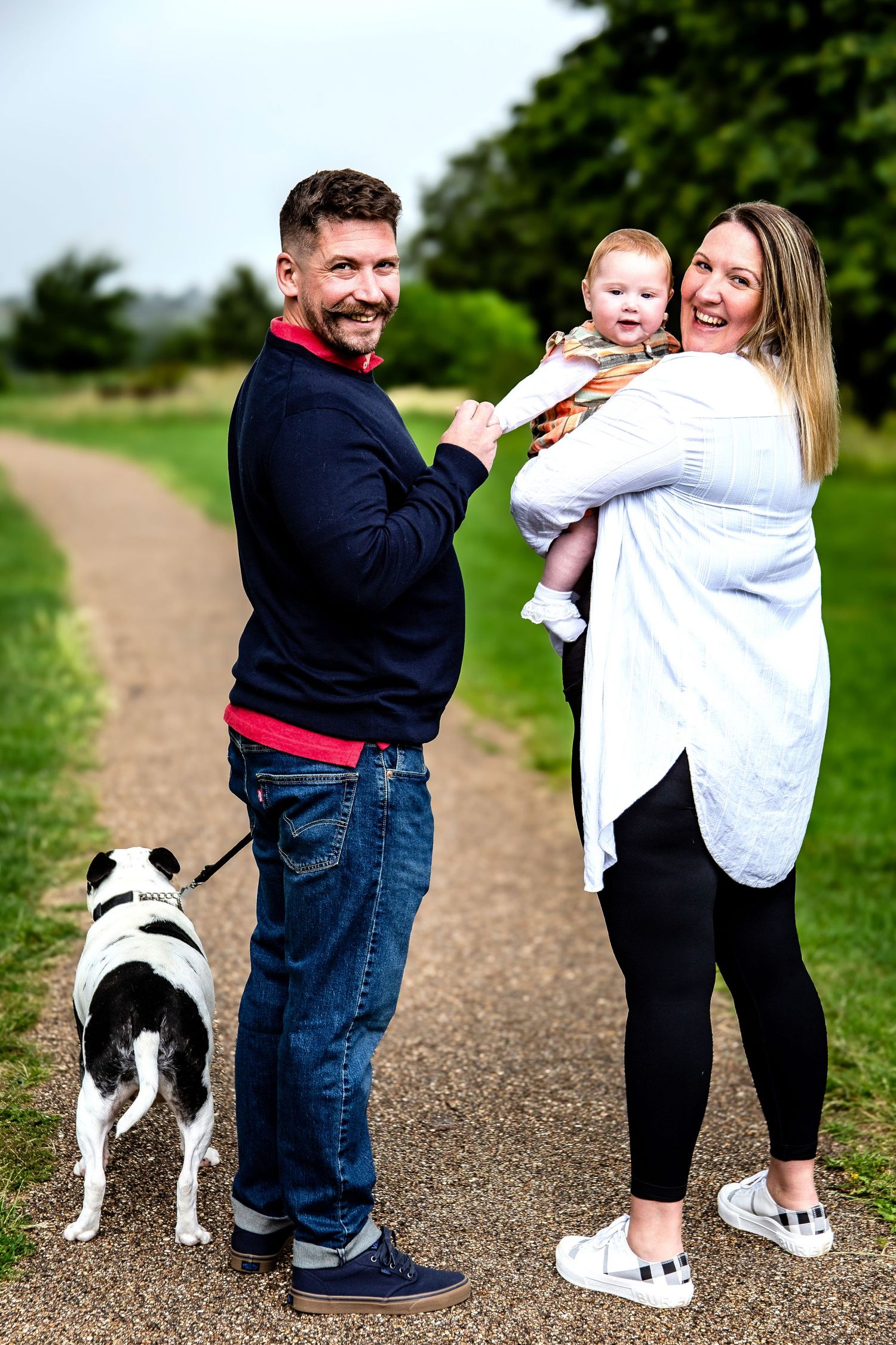 Smiling couple with baby and black and white dog posing on a park path outdoors.