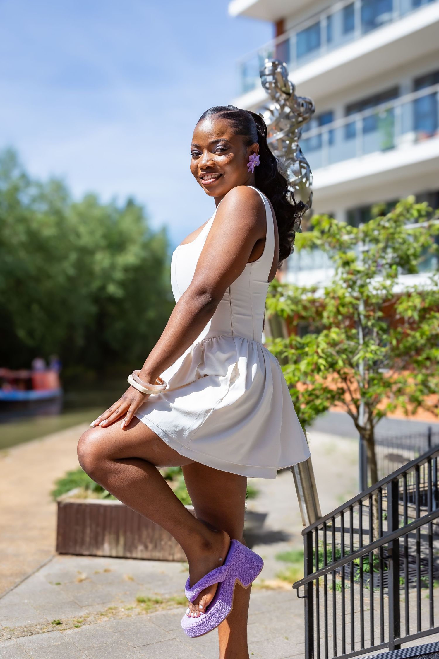 Smiling young woman in white mini dress and purple sandals posing on outdoor steps near a canal.