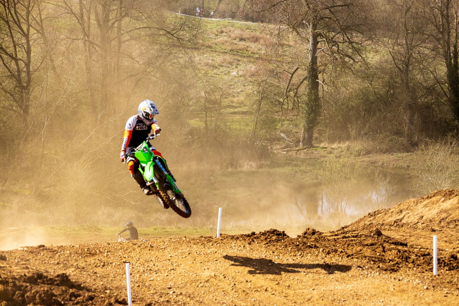 Motocross rider on green dirt bike catches air on a dusty track surrounded by trees.