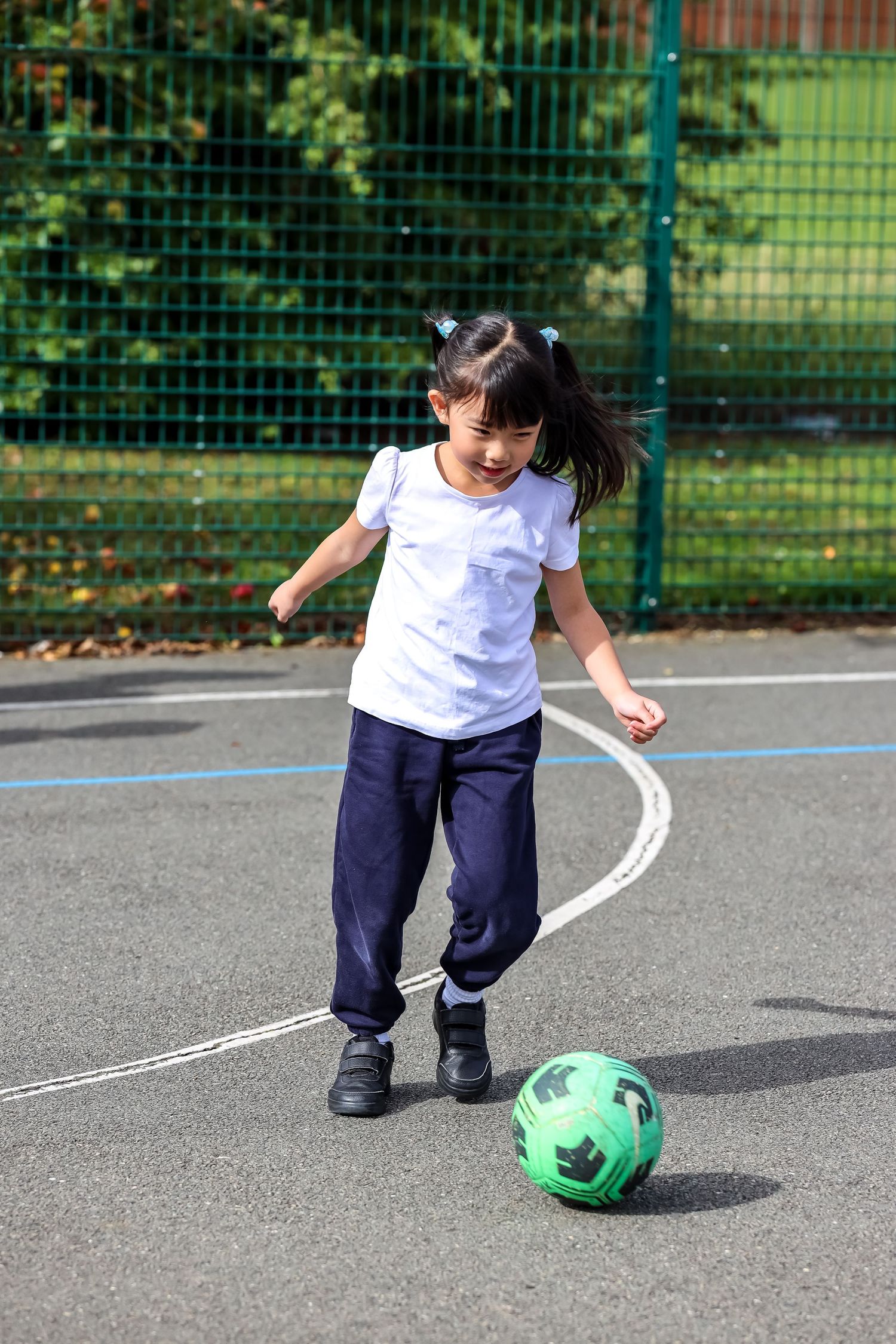 Young girl in school PE kit kicking a green football on an outdoor playground.