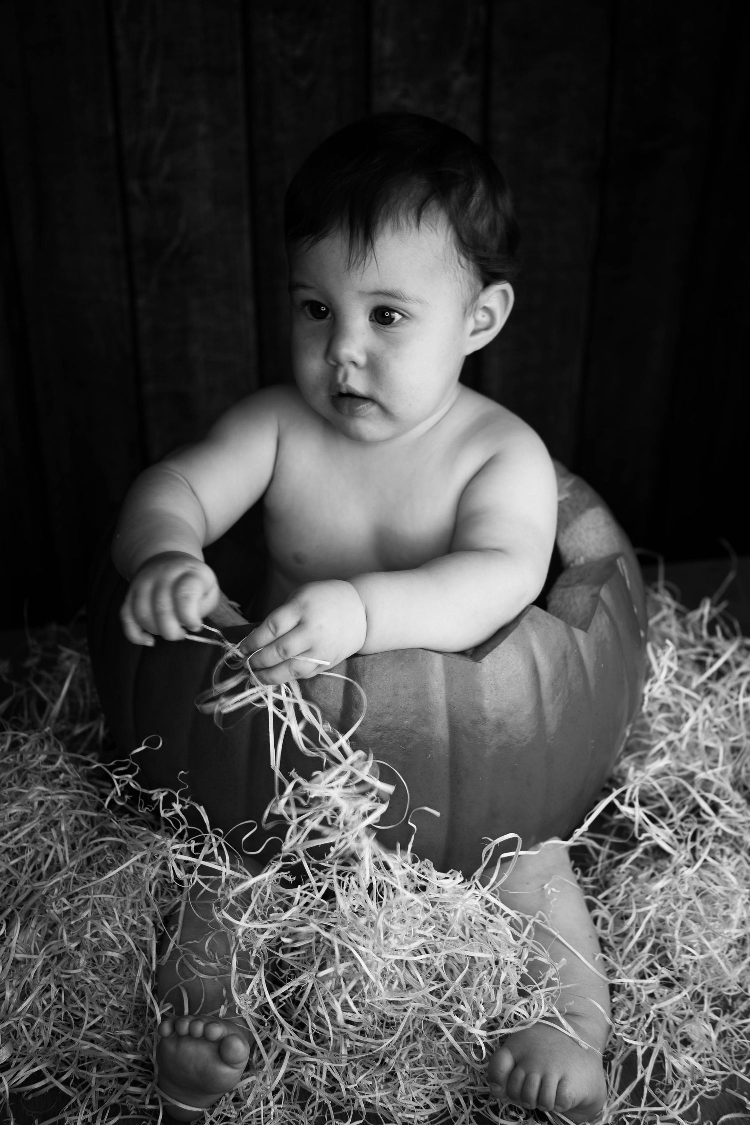 Black and white photo of a baby sitting inside a pumpkin surrounded by straw in a rustic setting.