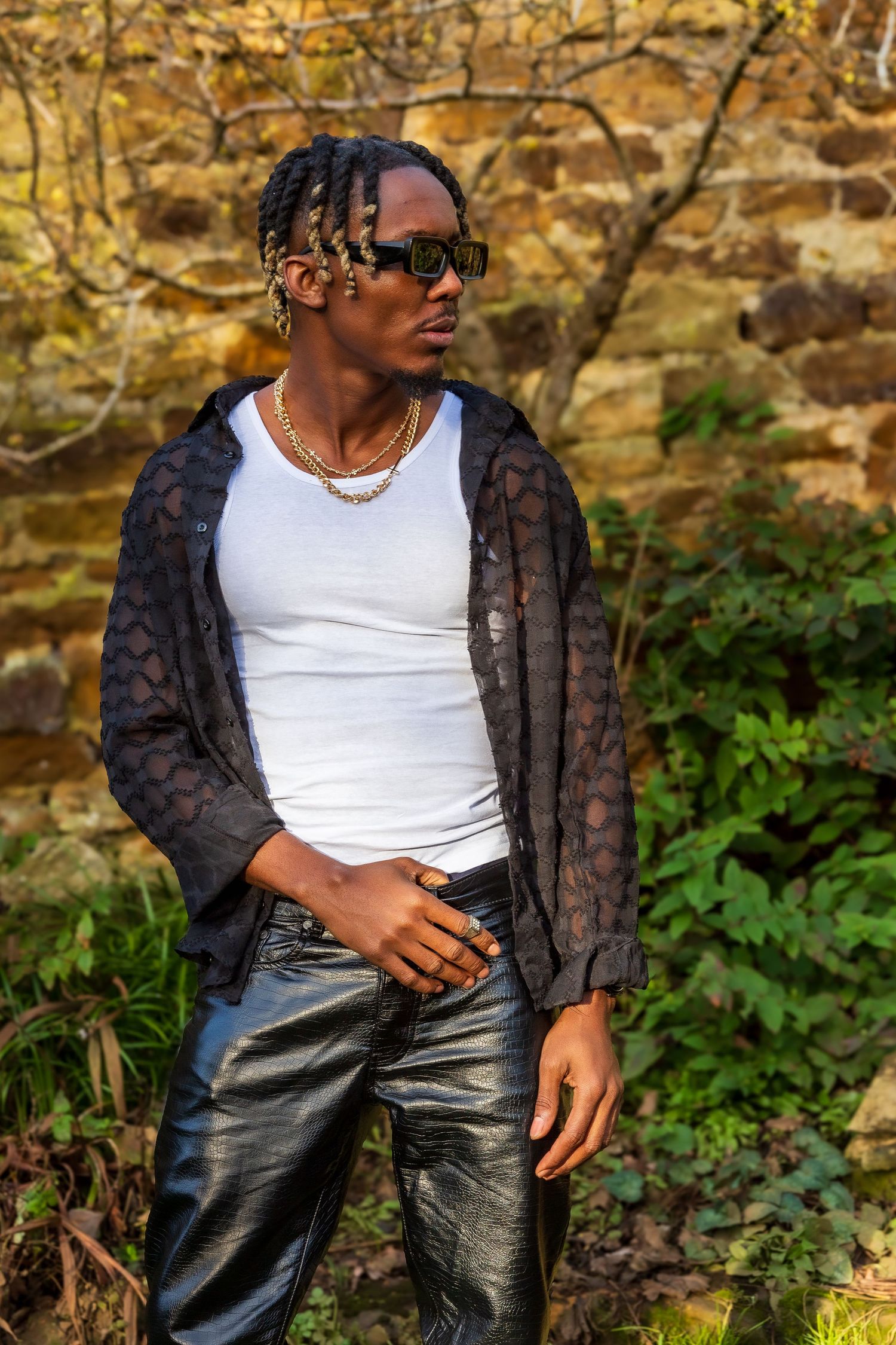 Stylish man modelling with braids wearing sunglasses, white tank top, and black leather pants outdoors.