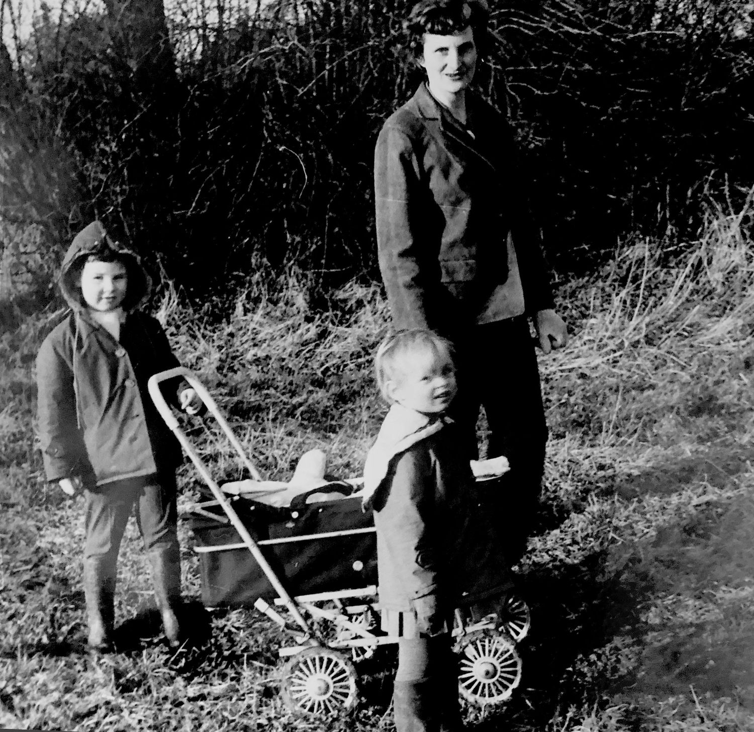 Vintage black and white photo of a woman with two young children and a pram outdoors in a wooded area.